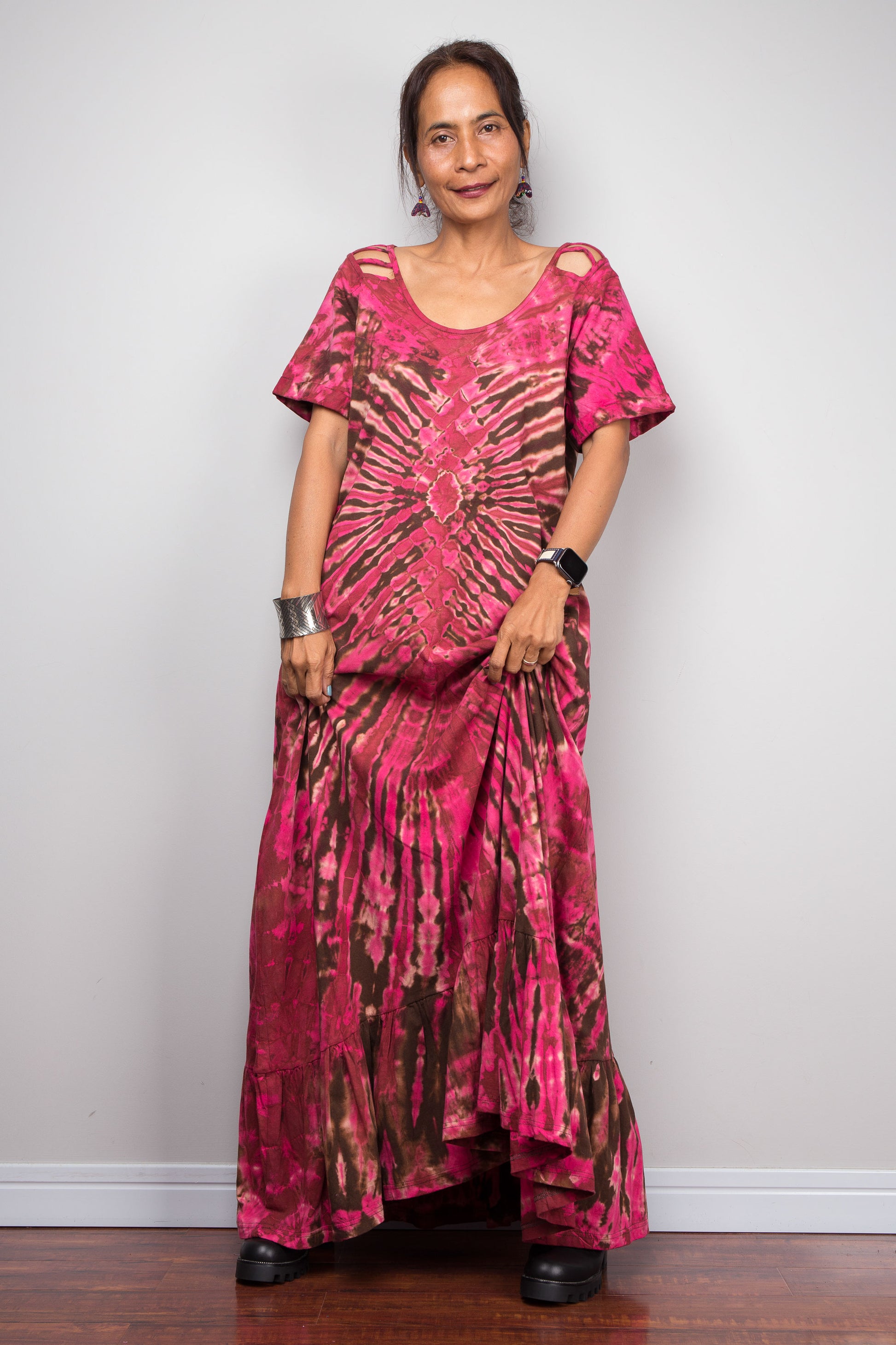 Front-facing view of a petite model wearing the Nuichan pink and brown tie dye maxi dress with hands in side pockets, showcasing the bold hot pink and chocolate brown starburst pattern, scoop neckline, short slashed sleeves.