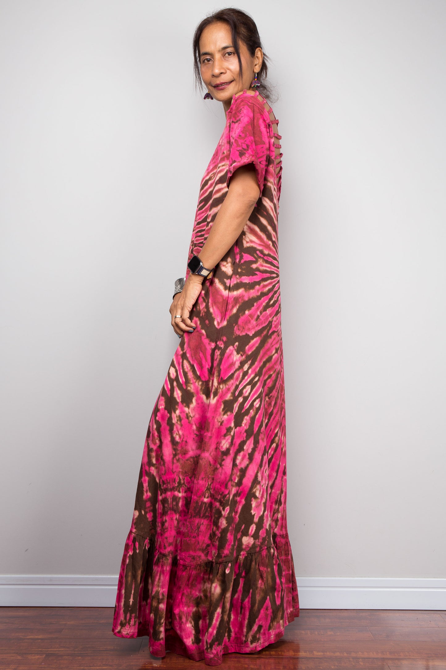 Side view of a model in the Nuichan pink tie dye maxi dress with hands on hips, highlighting the dramatic starburst radiating from a central point, relaxed A-line silhouette, cut-out shoulder details, and floor-length hem.