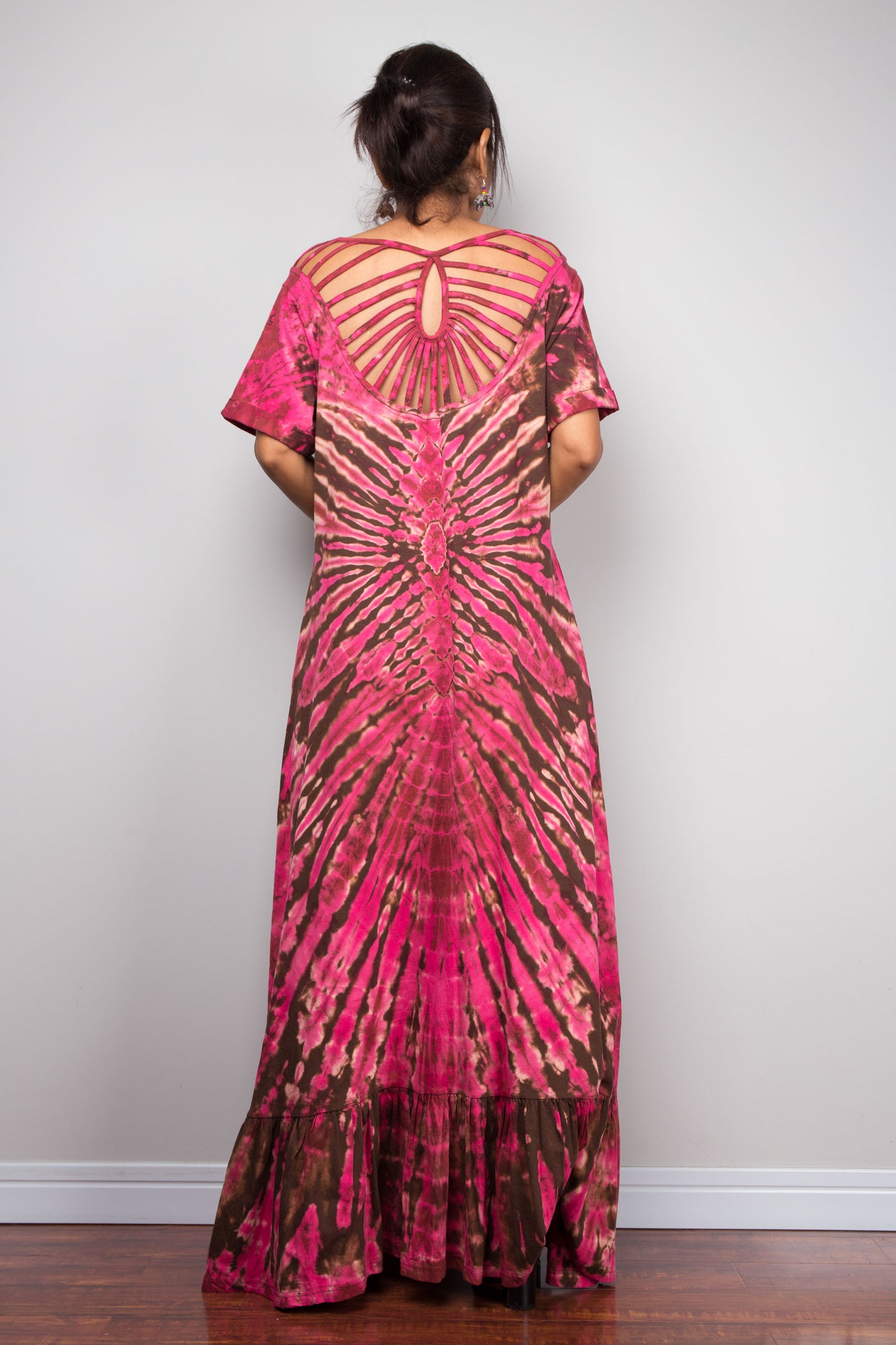 Rear view of a model wearing the Nuichan pink and brown tie dye maxi dress, showcasing the dramatic cage-style cut-out sunburst back design with fabric strips radiating from a central keyhole at the nape of the neck, and the full flowing A-line skirt.