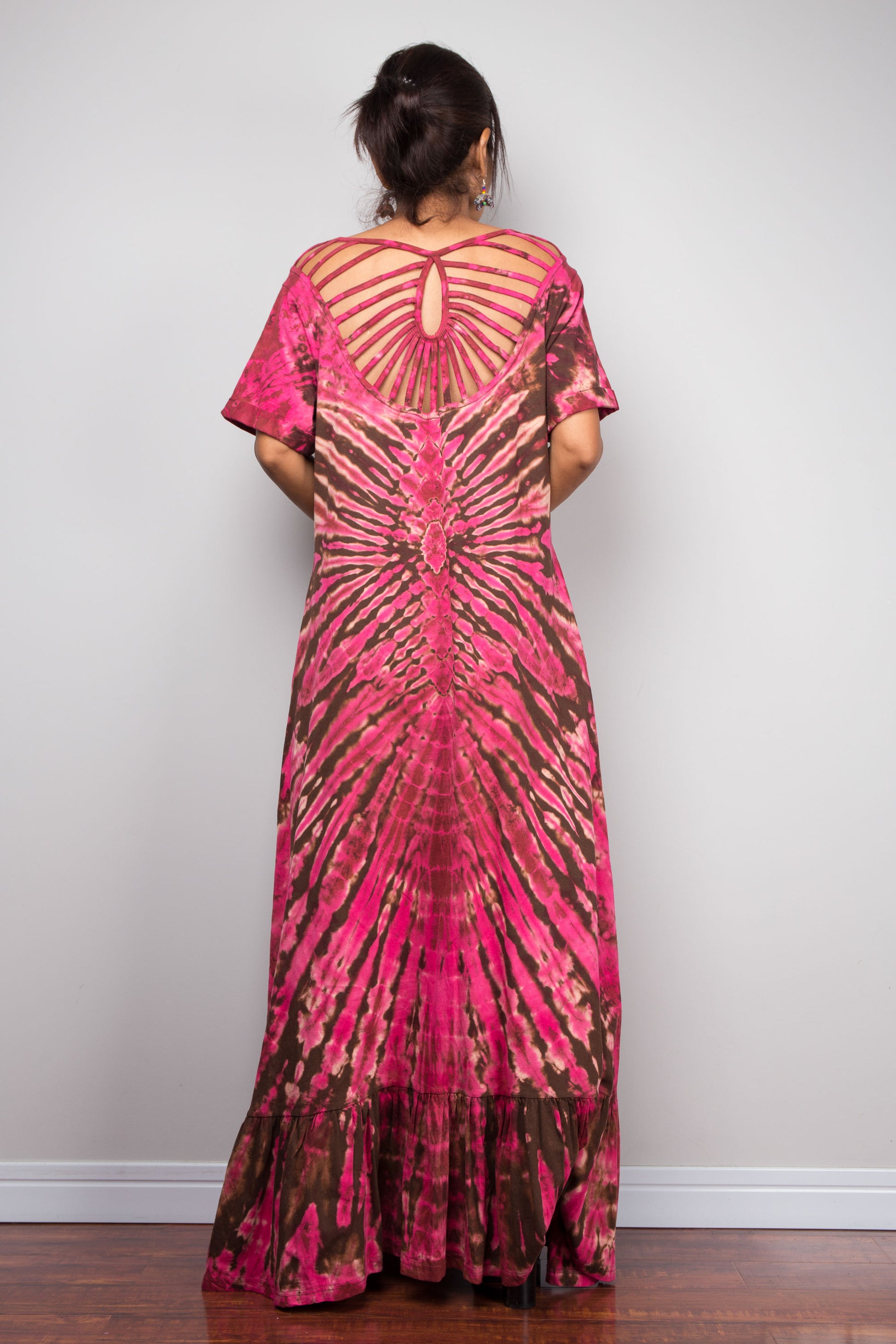 Rear view of a model wearing the Nuichan pink and brown tie dye maxi dress, showcasing the dramatic cage-style cut-out sunburst back design with fabric strips radiating from a central keyhole at the nape of the neck, and the full flowing A-line skirt.