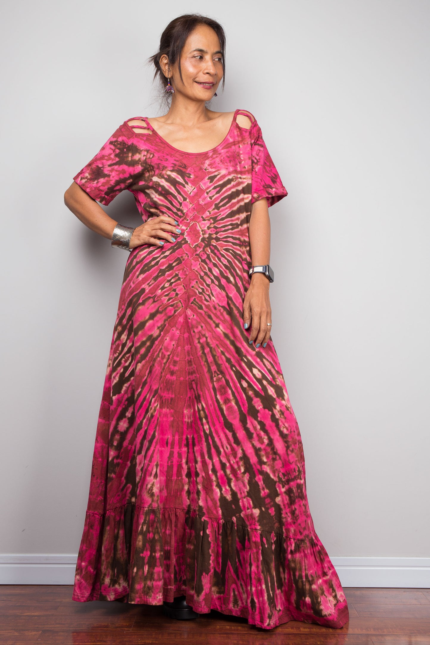 Three-quarter front view of a petite female model in the Nuichan pink tie dye maxi dress with hand on hip, highlighting the dramatic starburst radiating from a central diamond point, relaxed A-line silhouette, cut-out shoulder details, and floor-length hem.