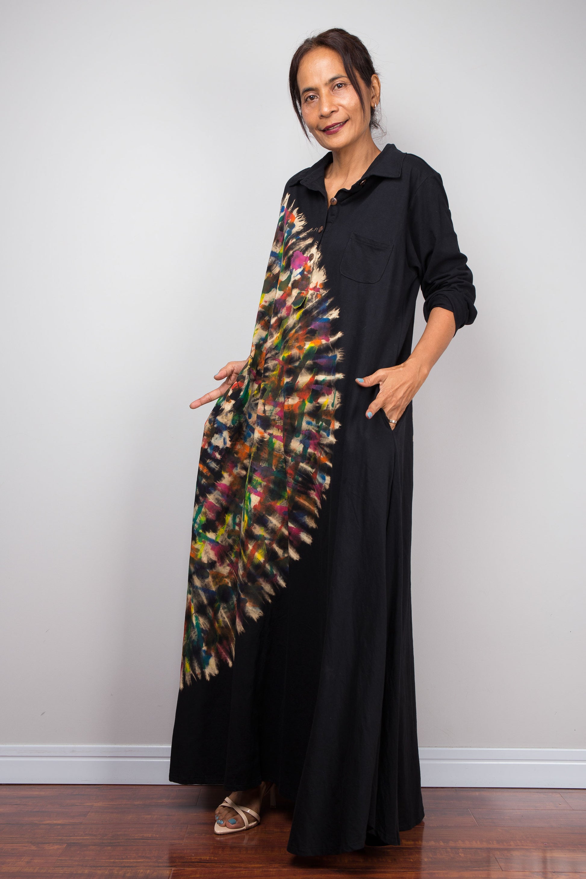 Side view of woman in black long-sleeve tie dye maxi dress. An asymmetric, vibrant hand-dyed multi-color tie-dye sunburst sweeps diagonally across the front. She has hands in side pockets.