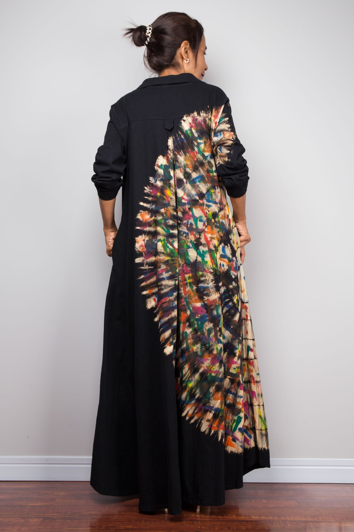 Back view of woman in long black long-sleeve maxi shirt dress. An asymmetric, vibrant hand-dyed multi-color tie-dye sunburst sweeps diagonally across the front. She has hands in side pockets.