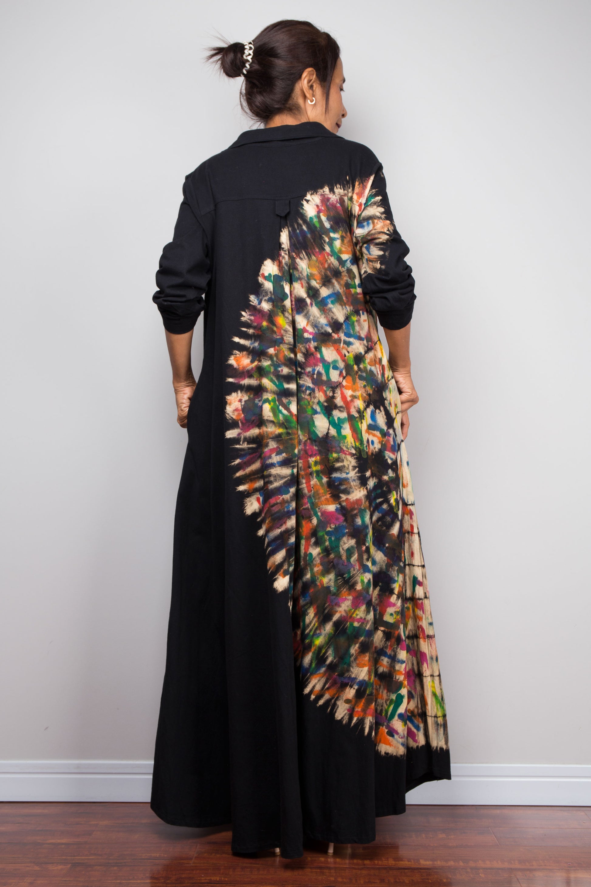 Back view of woman in long black long-sleeve maxi shirt dress. An asymmetric, vibrant hand-dyed multi-color tie-dye sunburst sweeps diagonally across the front. She has hands in side pockets.