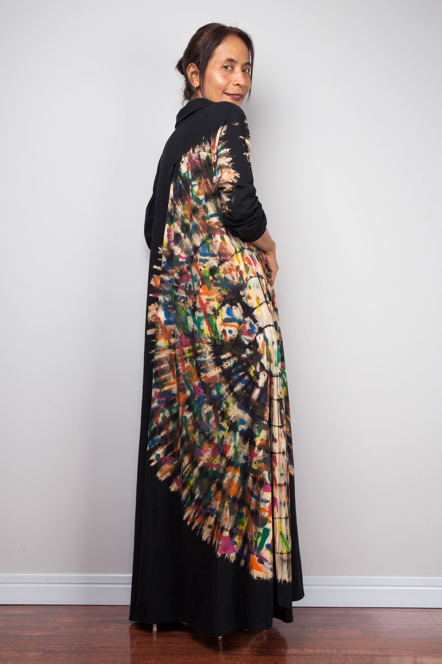 Back view of a Woman wearing a long, colorful patterned tie dye maxi dress against a plain background