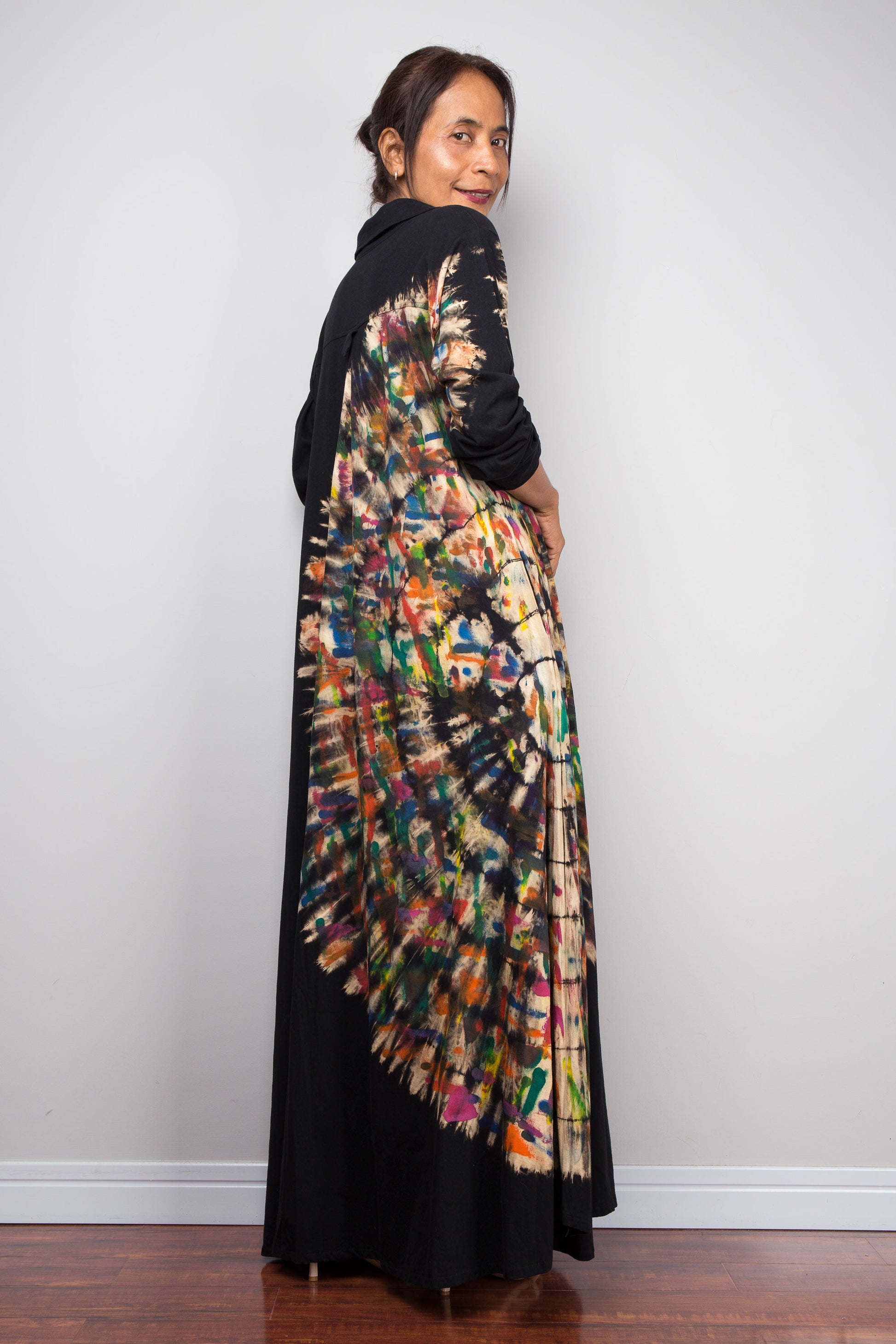 Back view of a Woman wearing a long, colorful patterned tie dye maxi dress against a plain background