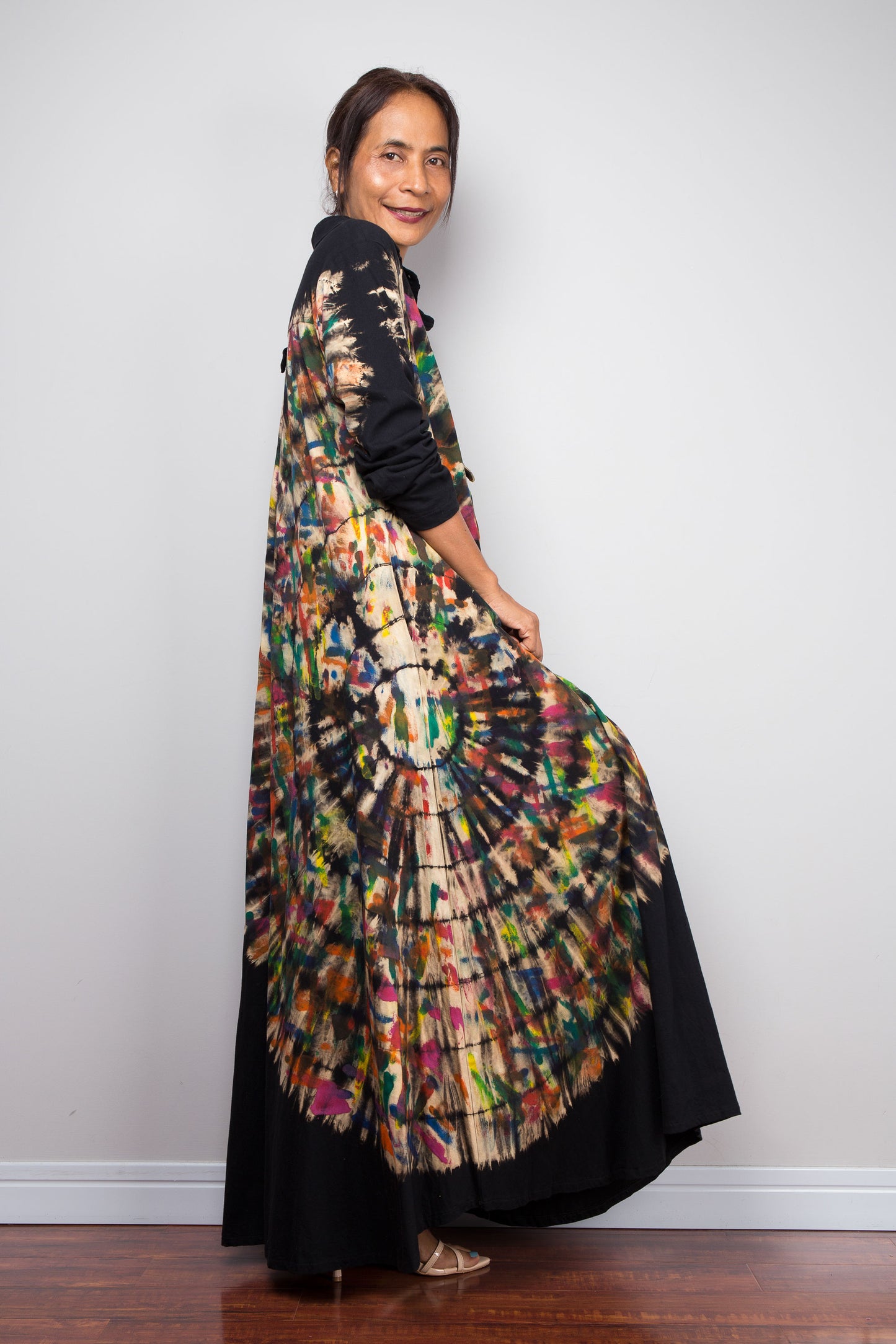 Side view of woman in black long-sleeve maxi shirt dress. An asymmetric, vibrant hand-dyed multi-color tie-dye sunburst sweeps diagonally across the front. 