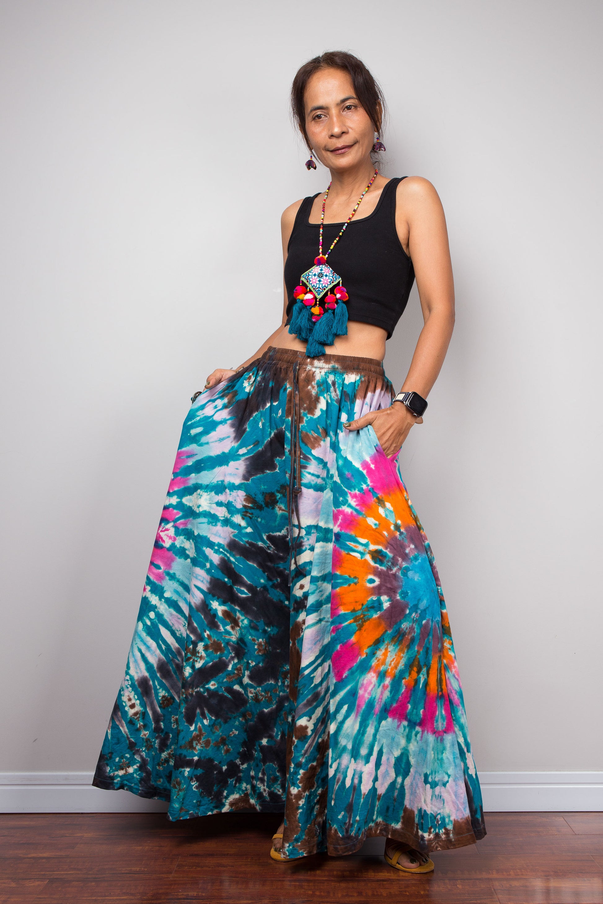 Three-quarter front view of a model wearing the Nuichan multicolour tie dye wide leg pants, highlighting the smocked chocolate brown elastic waistband with adjustable drawstring, inseam pockets and the dramatic floor-grazing A-line drape of the wide cotton legs