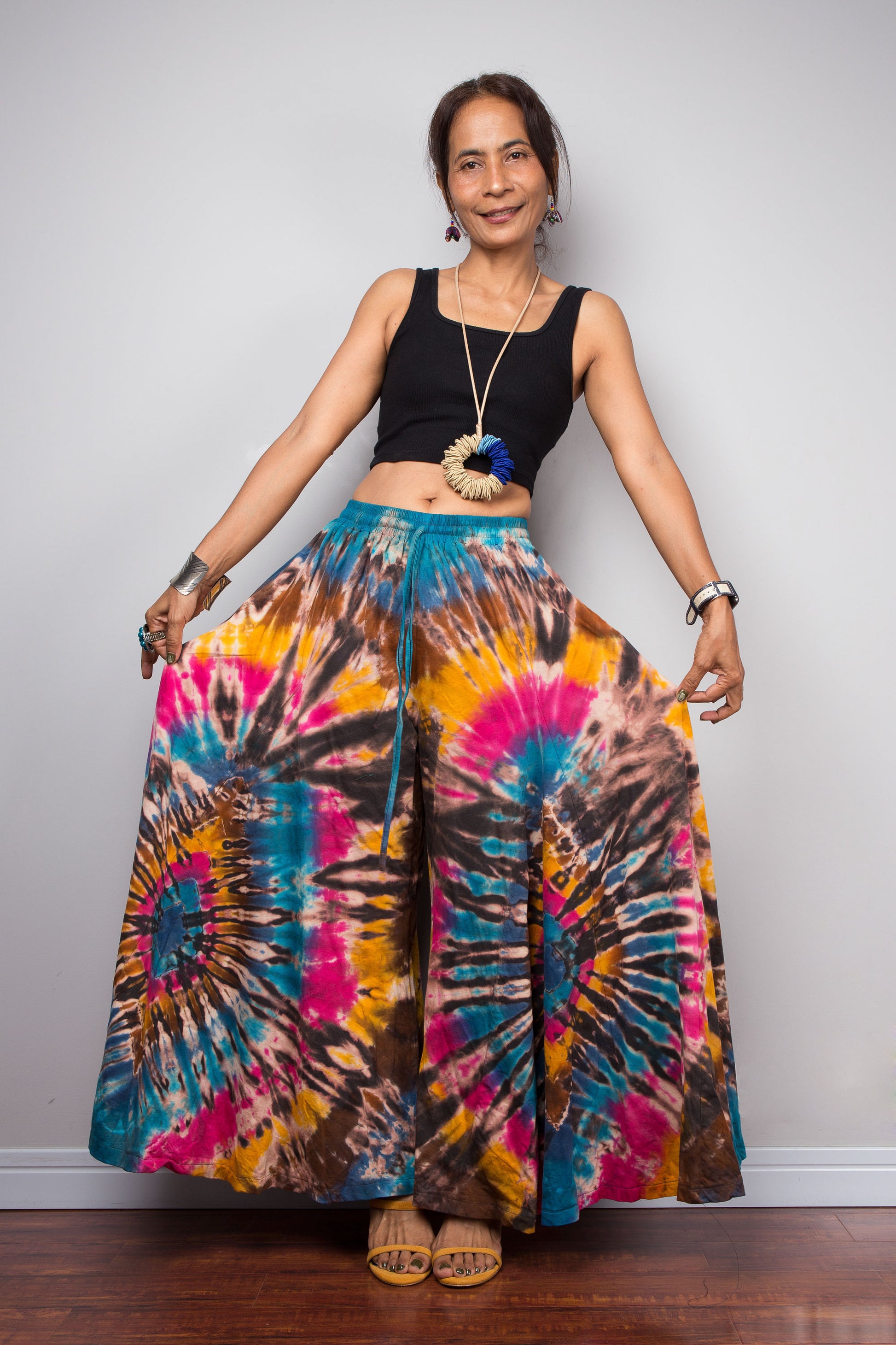 Front view of a model wearing the Nuichan turquoise, fuchsia and golden marigold tie dye wide leg palazzo pants in a dramatic spiral hand-dyed pattern, styled high-waisted with a black crop top, showcasing the flowing ultra-wide leg silhouette, vibrant artisan tie dye design, and turquoise smocked elastic waistband with adjustable drawstring.