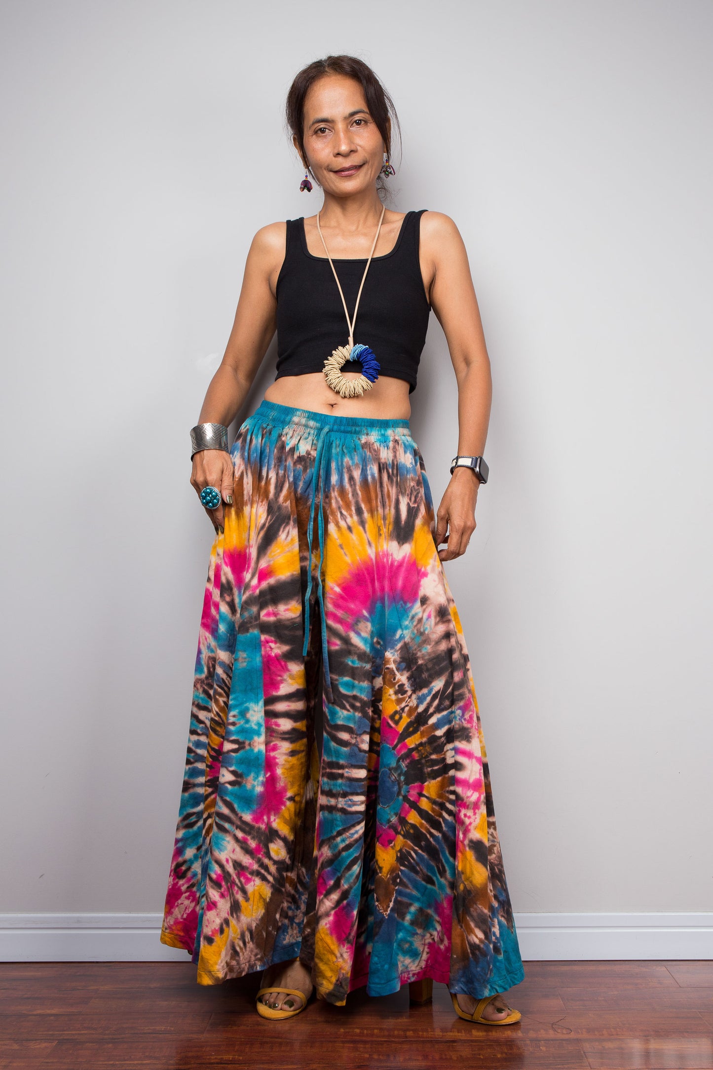 Front view of a petite model wearing the Nuichan turquoise, fuchsia and golden marigold tie dye wide leg palazzo pants, demonstrating the flattering high-waisted fit, full flowing silhouette, handcrafted spiral tie dye design, and elegant drape on a petite frame.