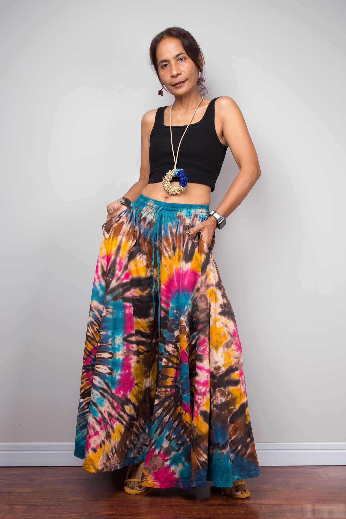 Lifestyle portrait of a model wearing the Nuichan artisan tie dye wide leg palazzo pants paired with a black crop top and handcrafted accessories, showcasing the inseam pockets, flowing bohemian silhouette, and effortless resort-inspired style.