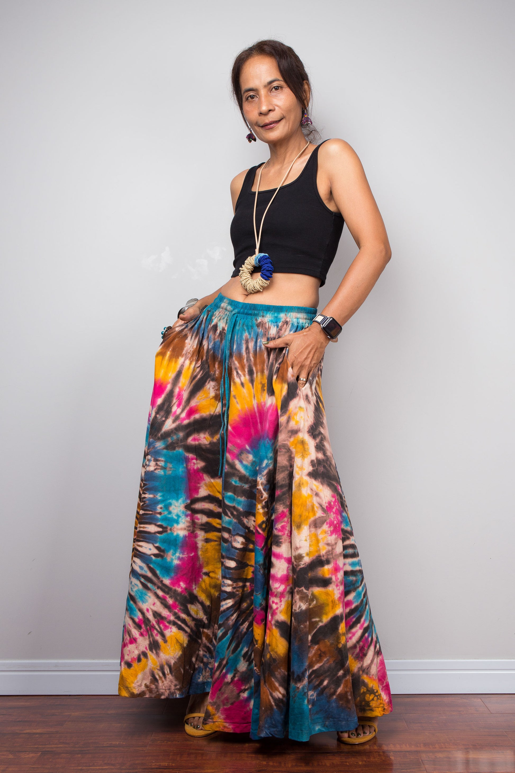 Three quarter view of a petite model wearing the Nuichan turquoise, fuchsia and golden marigold tie dye wide leg palazzo pants, demonstrating the flattering high-waisted fit, inseam pockets, full flowing silhouette, handcrafted spiral tie dye design, and elegant drape on a petite frame.