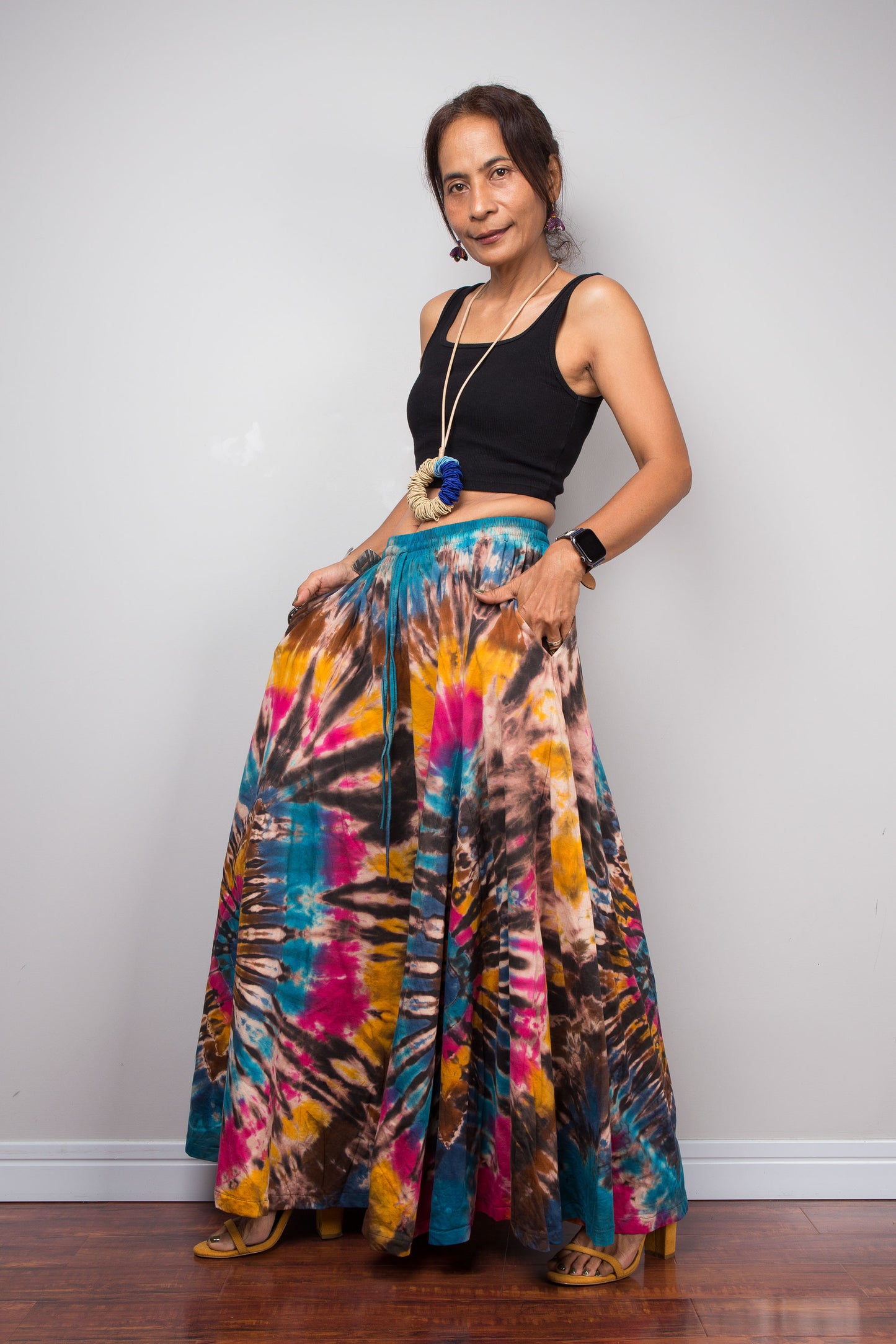 Three-quarter front view of a model wearing the Nuichan artisan tie dye wide leg palazzo pants featuring vibrant turquoise, fuchsia, golden marigold and chocolate brown spiral patterns, emphasizing the graceful drape, inseam pockets, flowing movement, handcrafted dye technique, and comfortable high-rise waist.