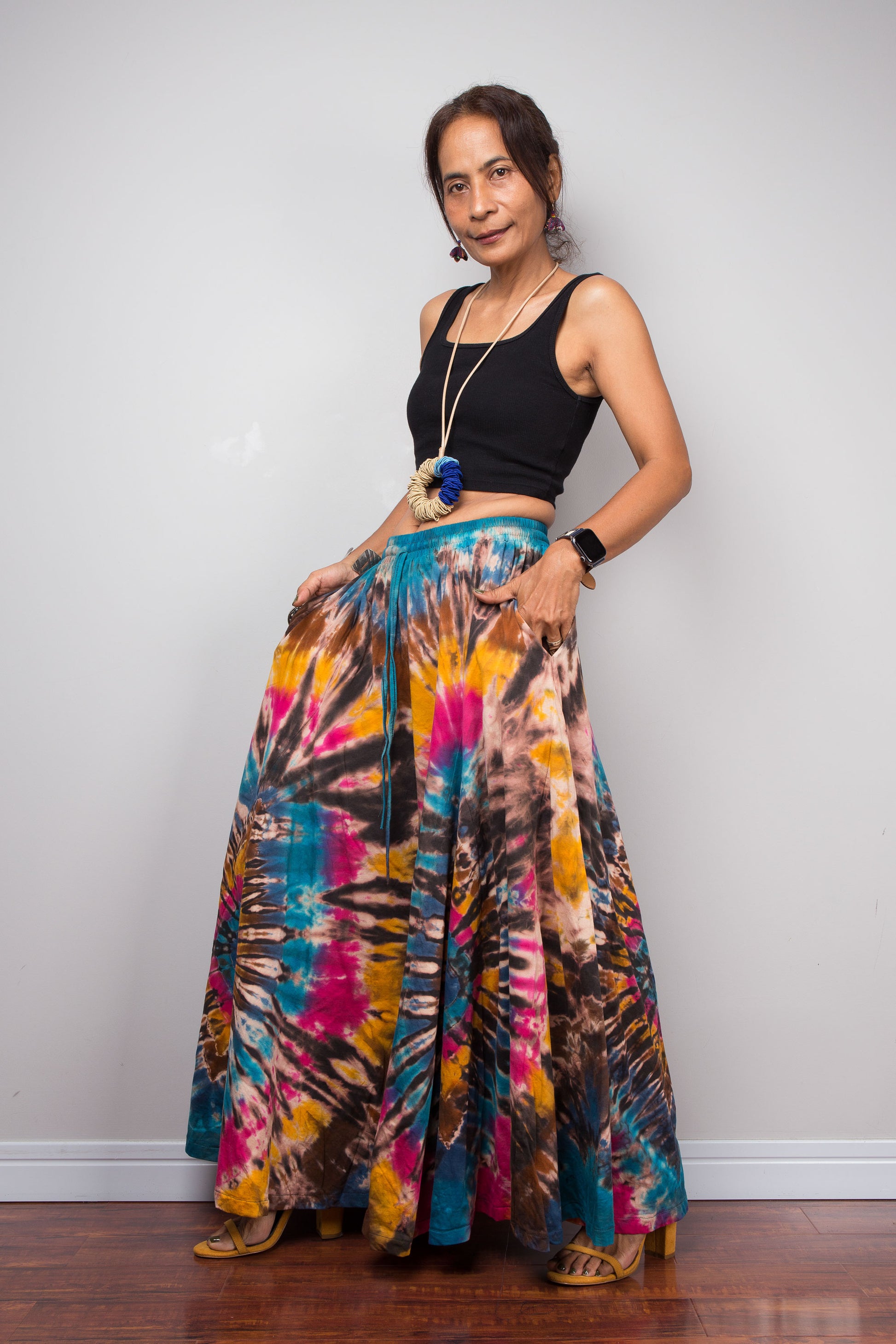 Three-quarter front view of a model wearing the Nuichan artisan tie dye wide leg palazzo pants featuring vibrant turquoise, fuchsia, golden marigold and chocolate brown spiral patterns, emphasizing the graceful drape, inseam pockets, flowing movement, handcrafted dye technique, and comfortable high-rise waist.
