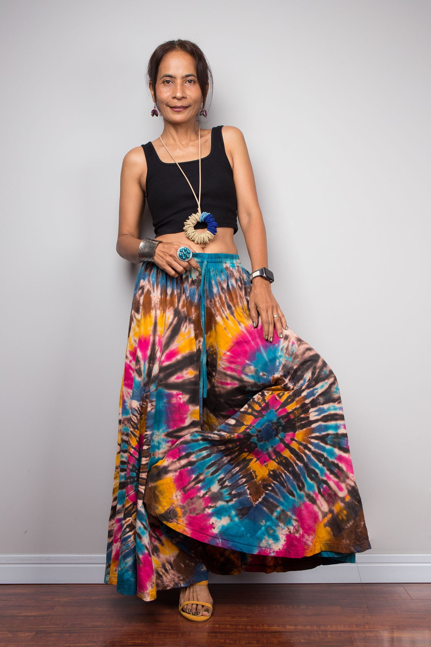 Front view of a petite female model wearing the Nuichan turquoise, fuchsia and golden marigold tie dye wide leg palazzo pants, demonstrating the flattering high-waisted fit, full flowing silhouette, handcrafted spiral tie dye design, and elegant drape on a petite frame.