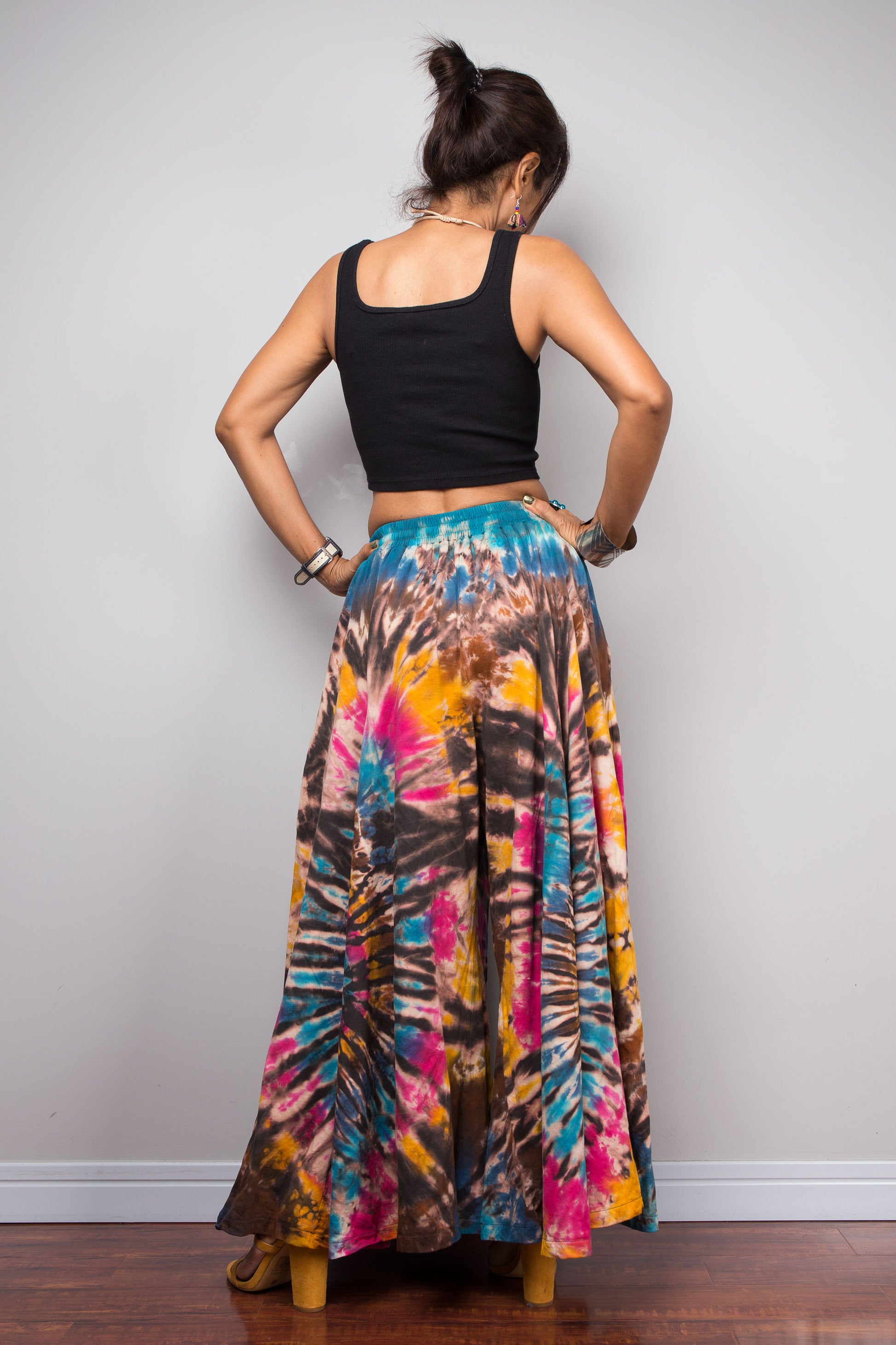 Rear view of a petite model wearing the Nuichan turquoise, fuchsia and golden marigold tie dye wide leg palazzo pants, highlighting the full sweeping palazzo silhouette, striking spiral tie dye artwork that continues across the back, comfortable smocked waistband, and relaxed bohemian fit.