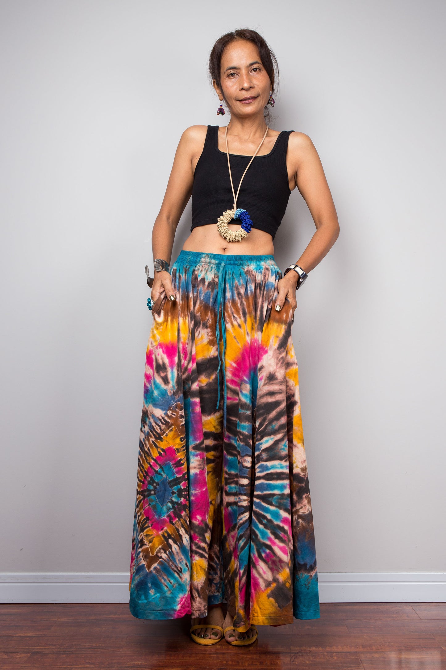 Front view of a petite female model wearing the Nuichan turquoise, fuchsia and golden marigold tie dye wide leg palazzo pants in a dramatic spiral hand-dyed pattern, styled high-waisted with a black crop top, showcasing the flowing ultra-wide leg silhouette, vibrant artisan tie dye design, inseam pockets and smocked elastic waistband with adjustable drawstring.