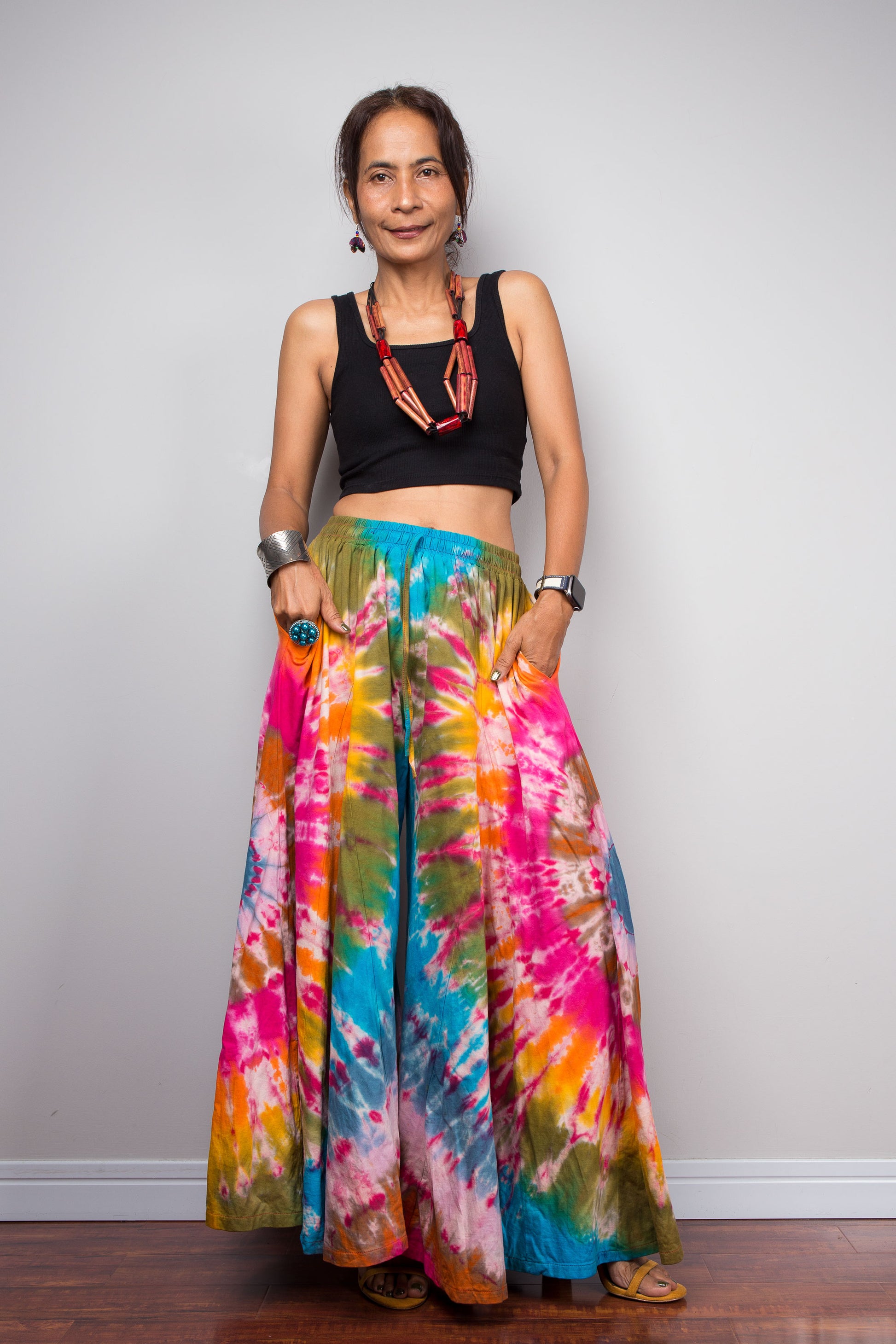 Front view of a petite model wearing the Nuichan rainbow carnival tie dye wide leg palazzo pants with hands in inseam pockets, showcasing the complex multi-point starburst hand-dyed pattern in hot pink magenta turquoise orange and olive, dramatic floor-length ultra-wide leg silhouette, and distinctive dual-tone smocked elastic waistband in turquoise and olive, styled with a black crop top