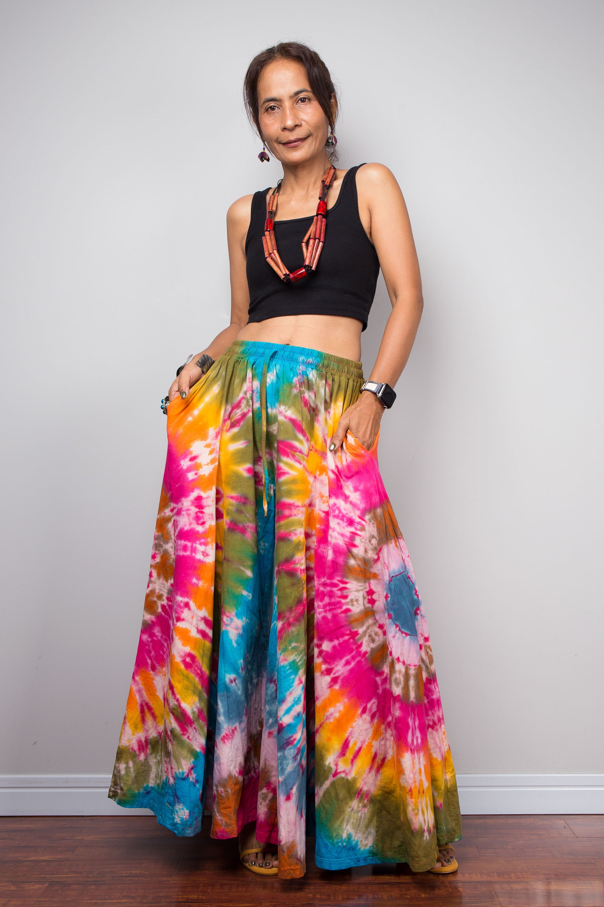 Front-facing view of a model wearing the Nuichan rainbow carnival tie dye wide leg palazzo pants with hands in inseam pockets, showcasing the complex multi-point starburst hand-dyed pattern in hot pink magenta turquoise orange and olive, dramatic floor-length ultra-wide leg silhouette, and distinctive dual-tone smocked elastic waistband in turquoise and olive, styled with a black crop top