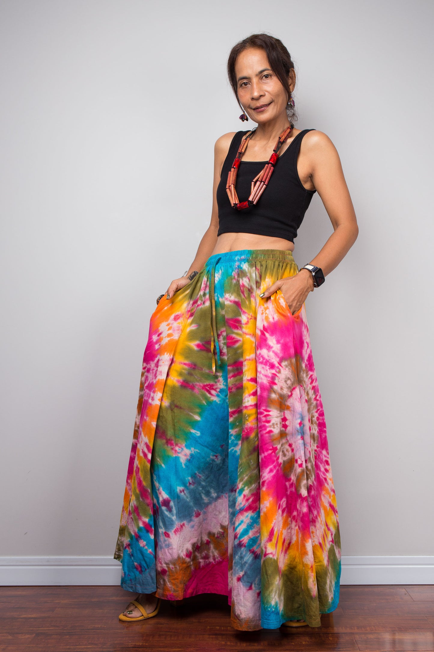 Side facing view of a model wearing the Nuichan rainbow carnival tie dye wide leg palazzo pants with hands in inseam pockets, showcasing the complex multi-point starburst hand-dyed pattern in hot pink magenta turquoise orange and olive, dramatic floor-length ultra-wide leg silhouette, and distinctive dual-tone smocked elastic waistband in turquoise and olive, styled with a black crop top