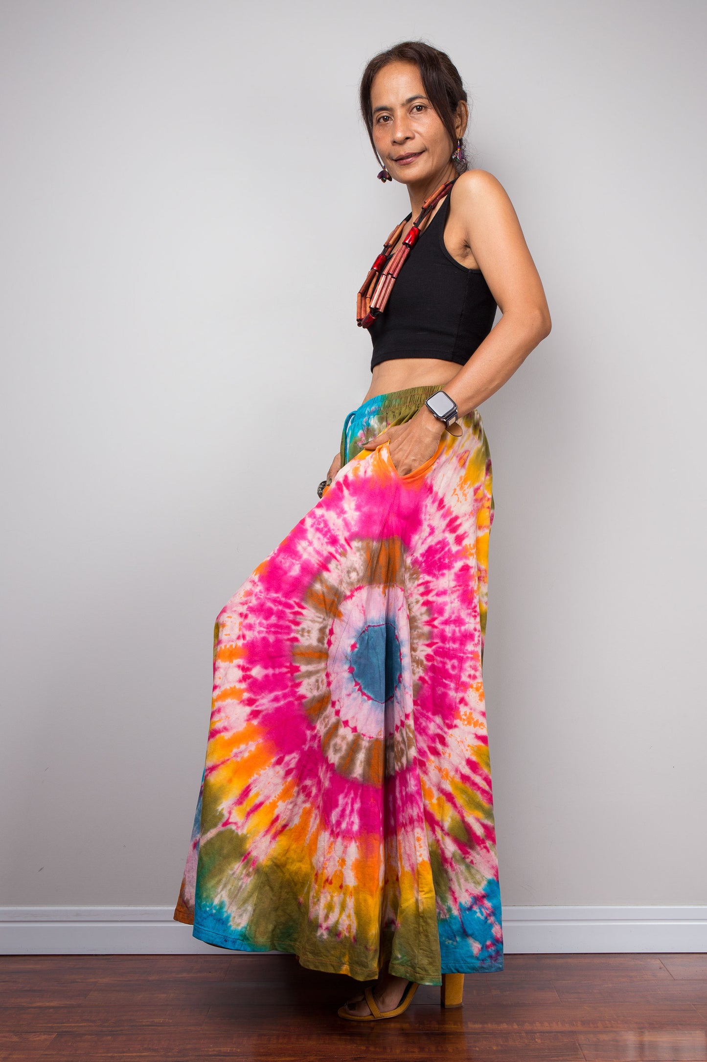 Side on view of a petite model wearing the Nuichan rainbow carnival tie dye wide leg palazzo pants with hands in inseam pockets, showcasing the complex multi-point starburst hand-dyed pattern in hot pink magenta turquoise orange and olive, dramatic floor-length ultra-wide leg silhouette, and distinctive dual-tone smocked elastic waistband in turquoise and olive, styled with a black crop top