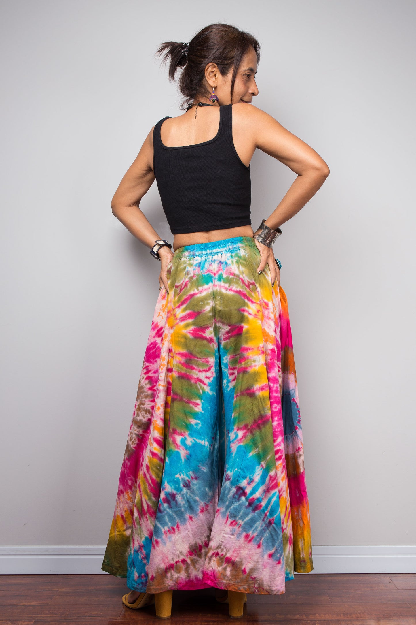 Rear view of a model wearing the Nuichan rainbow carnival tie dye wide leg palazzo pants with hands on hips, showcasing the complex multi-point starburst hand-dyed pattern in hot pink magenta turquoise orange and olive, dramatic floor-length ultra-wide leg silhouette, and distinctive dual-tone smocked elastic waistband in turquoise and olive, styled with a black crop top