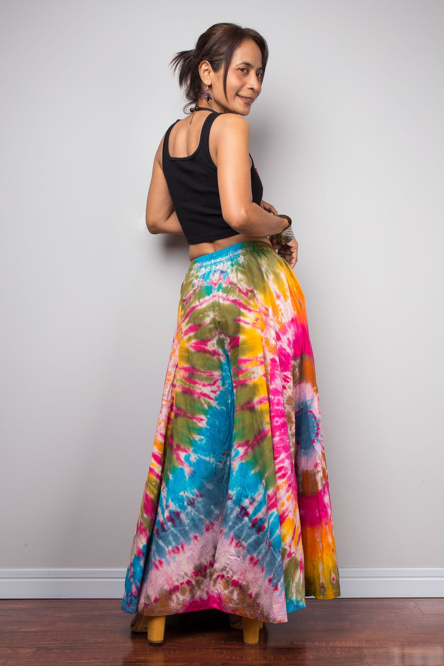 Back facing view of a model wearing the Nuichan rainbow carnival tie dye wide leg palazzo pants, showcasing the complex multi-point starburst hand-dyed pattern in hot pink magenta turquoise orange and olive, dramatic floor-length ultra-wide leg silhouette, and distinctive dual-tone smocked elastic waistband in turquoise and olive, styled with a black crop top