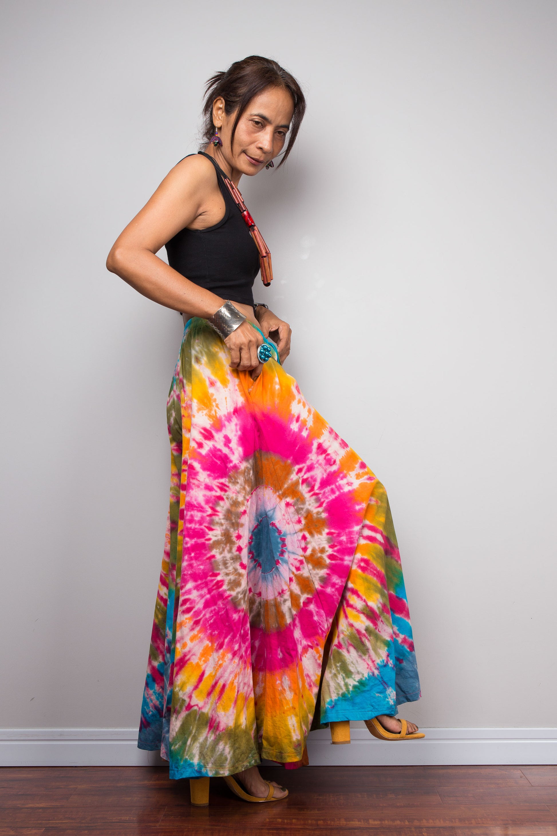 Side view of a model wearing the Nuichan rainbow carnival tie dye wide leg palazzo pants, showcasing the complex multi-point starburst hand-dyed pattern in hot pink magenta turquoise orange and olive, dramatic floor-length ultra-wide leg silhouette, and distinctive dual-tone smocked elastic waistband in turquoise and olive, styled with a black crop top