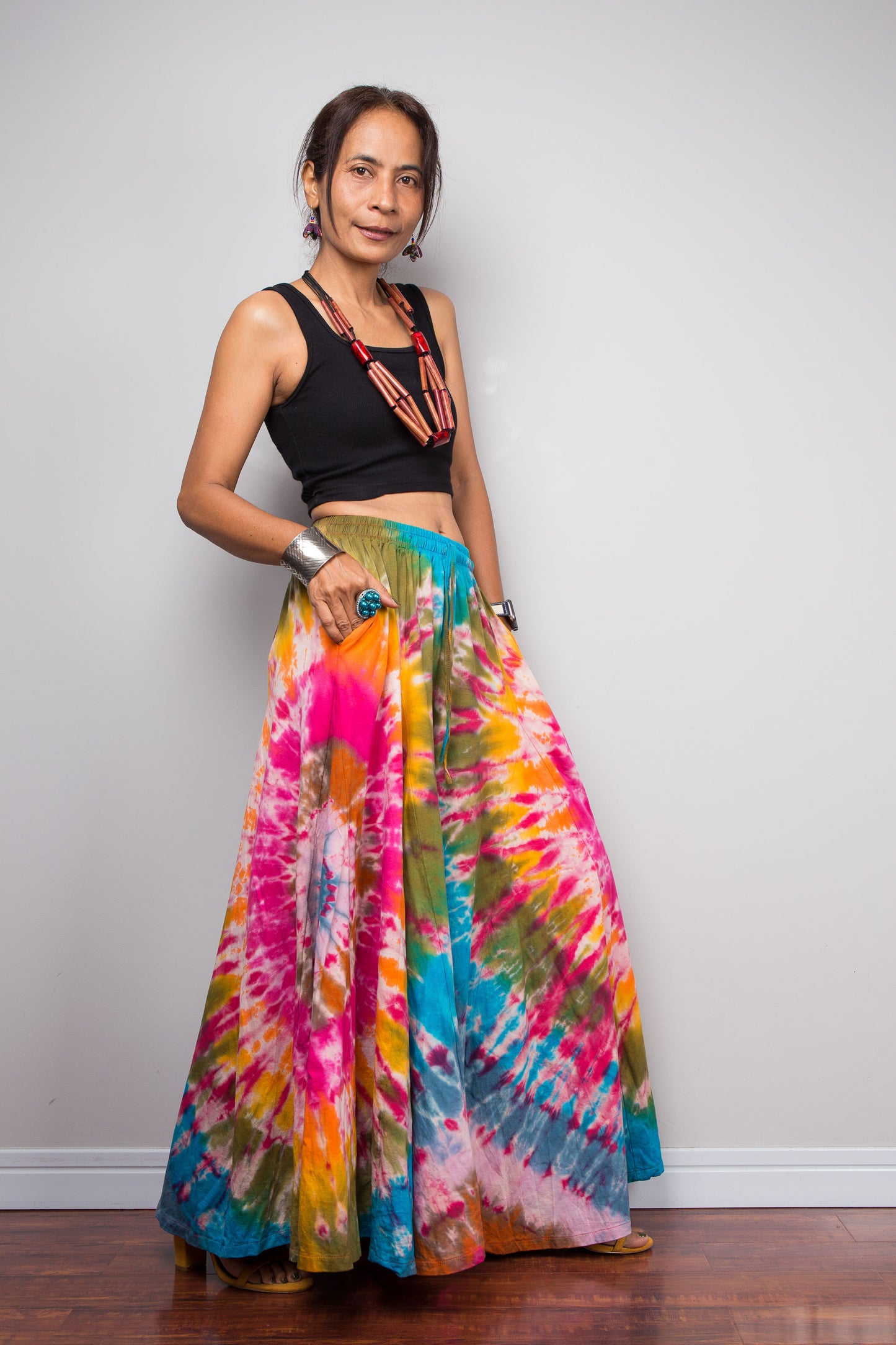 Three quarter view of a model wearing the Nuichan rainbow carnival tie dye wide leg palazzo pants with hands in inseam pockets, showcasing the complex multi-point starburst hand-dyed pattern in hot pink magenta turquoise orange and olive, dramatic floor-length ultra-wide leg silhouette, and distinctive dual-tone smocked elastic waistband in turquoise and olive, styled with a black crop top