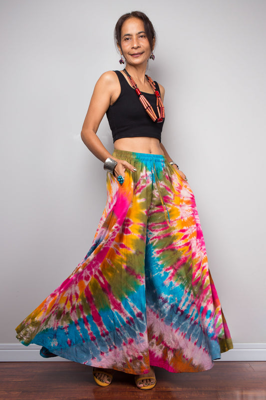 Dynamic view of a model wearing the Nuichan rainbow carnival tie dye wide leg palazzo pants with hands in inseam pockets, showcasing the complex multi-point starburst hand-dyed pattern in hot pink magenta turquoise orange and olive, dramatic floor-length ultra-wide leg silhouette, and distinctive dual-tone smocked elastic waistband in turquoise and olive, styled with a black crop top