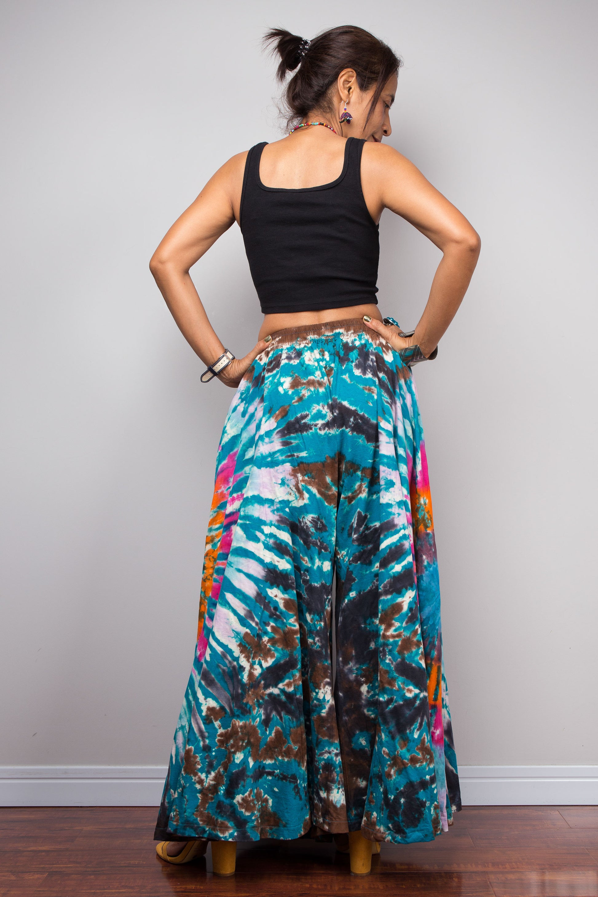 Rear view of a model wearing the Nuichan multicolour tie dye palazzo pants with hands on hips, displaying the full width of the ultra-wide leg silhouette, smocked waistband detail, and the continuation of the freeform teal pink and brown tie dye pattern across the back