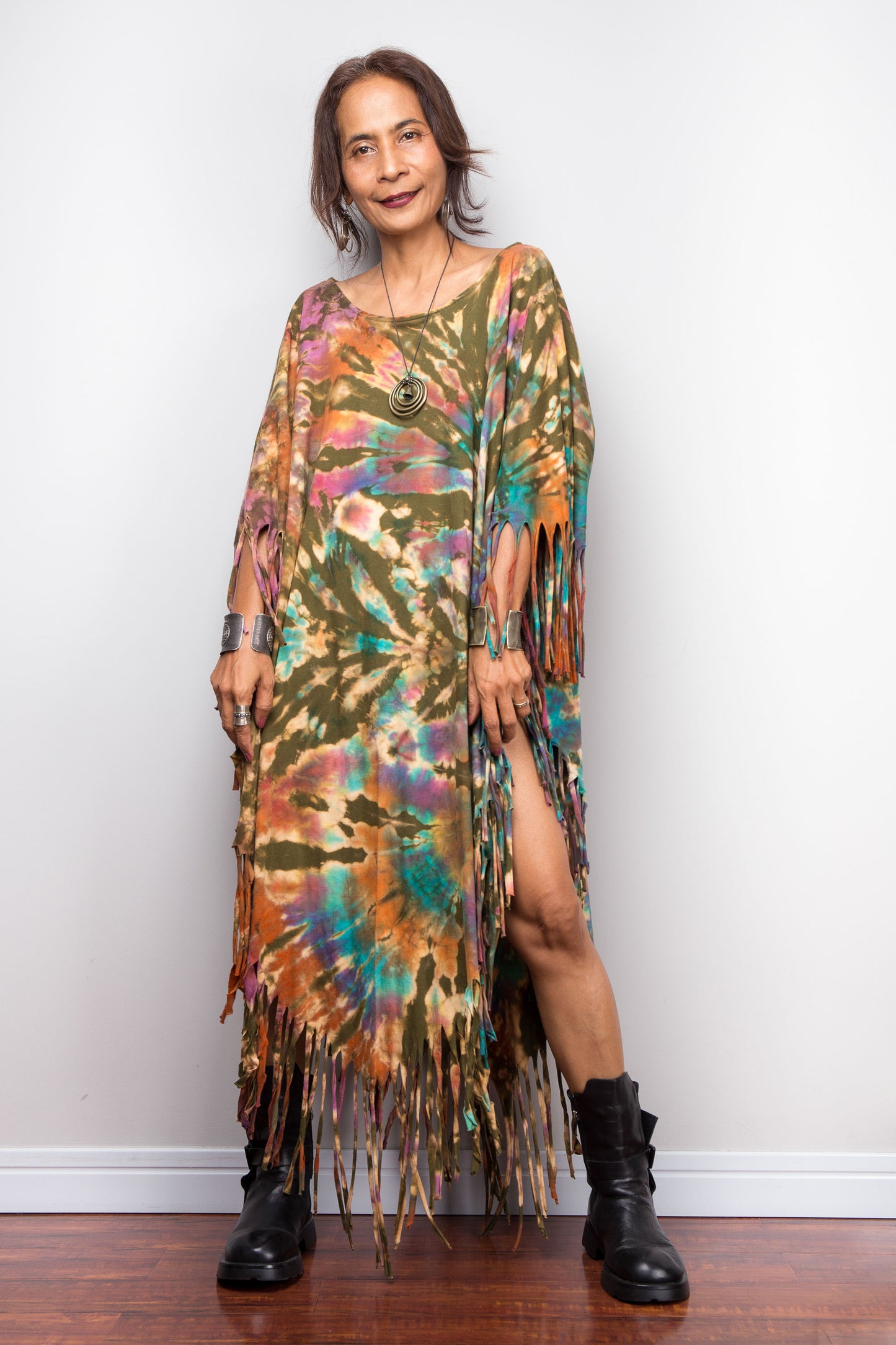 Woman wearing a colorful tie dye poncho dress with fringe standing against a white wall.