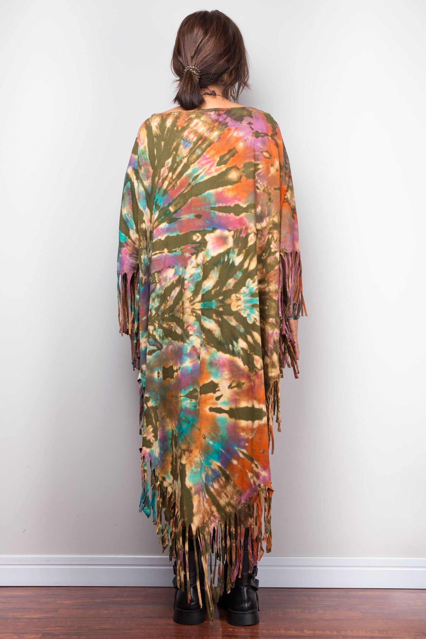 Person wearing a tie-dye poncho with fringe details against a white wall.