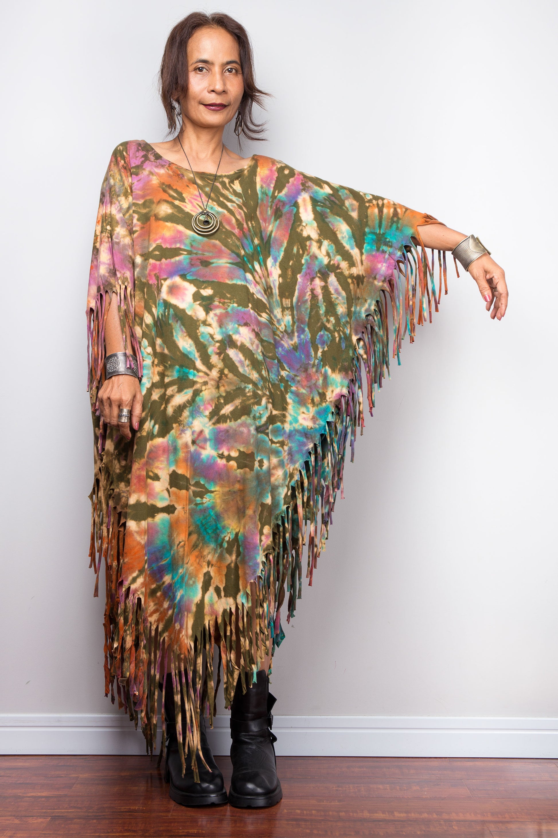 Woman wearing a colorful tie dye poncho with fringes against a white wall.