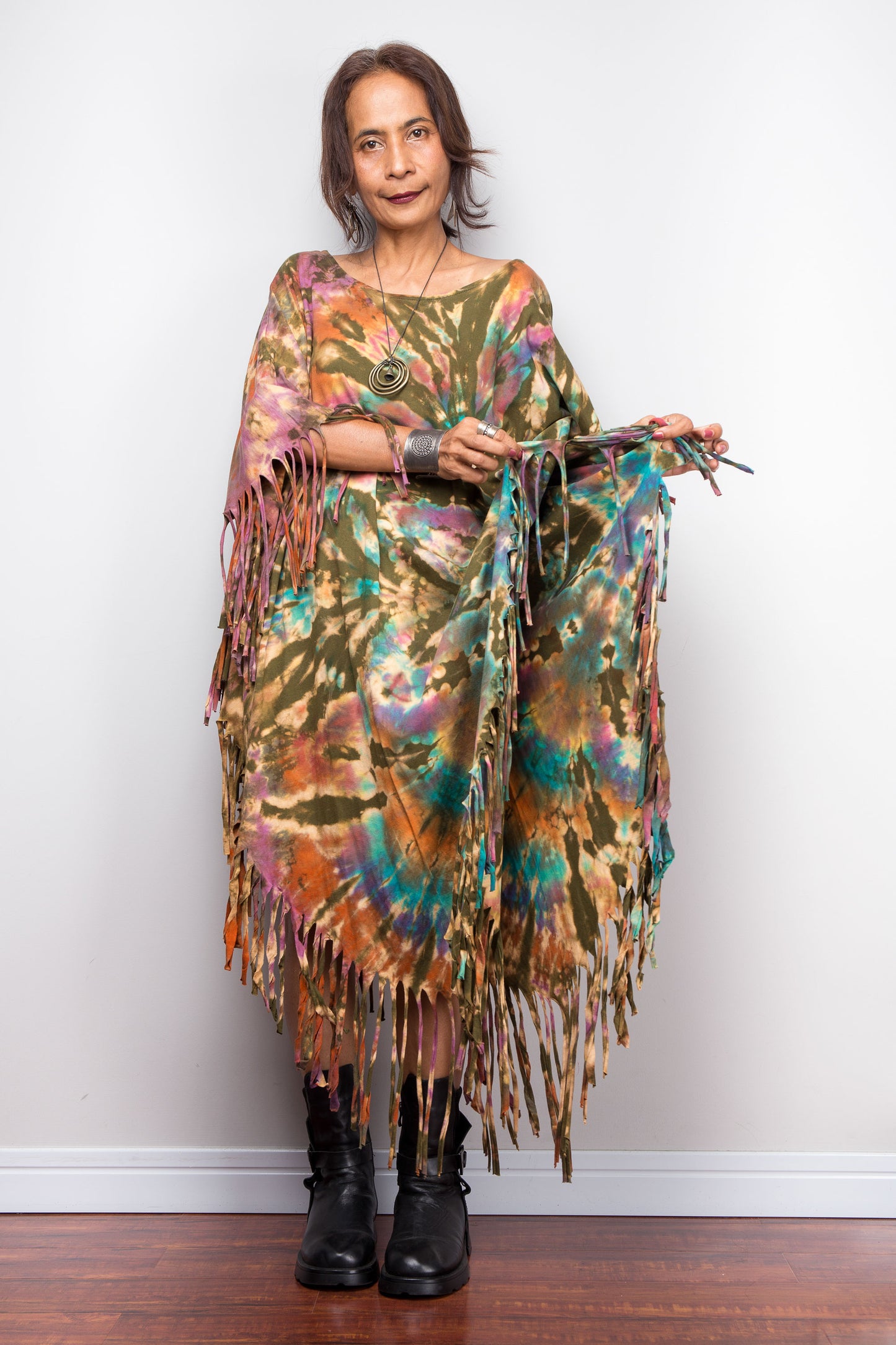 Woman wearing a colorful tie dye poncho kaftan dress with fringe against a white wall.