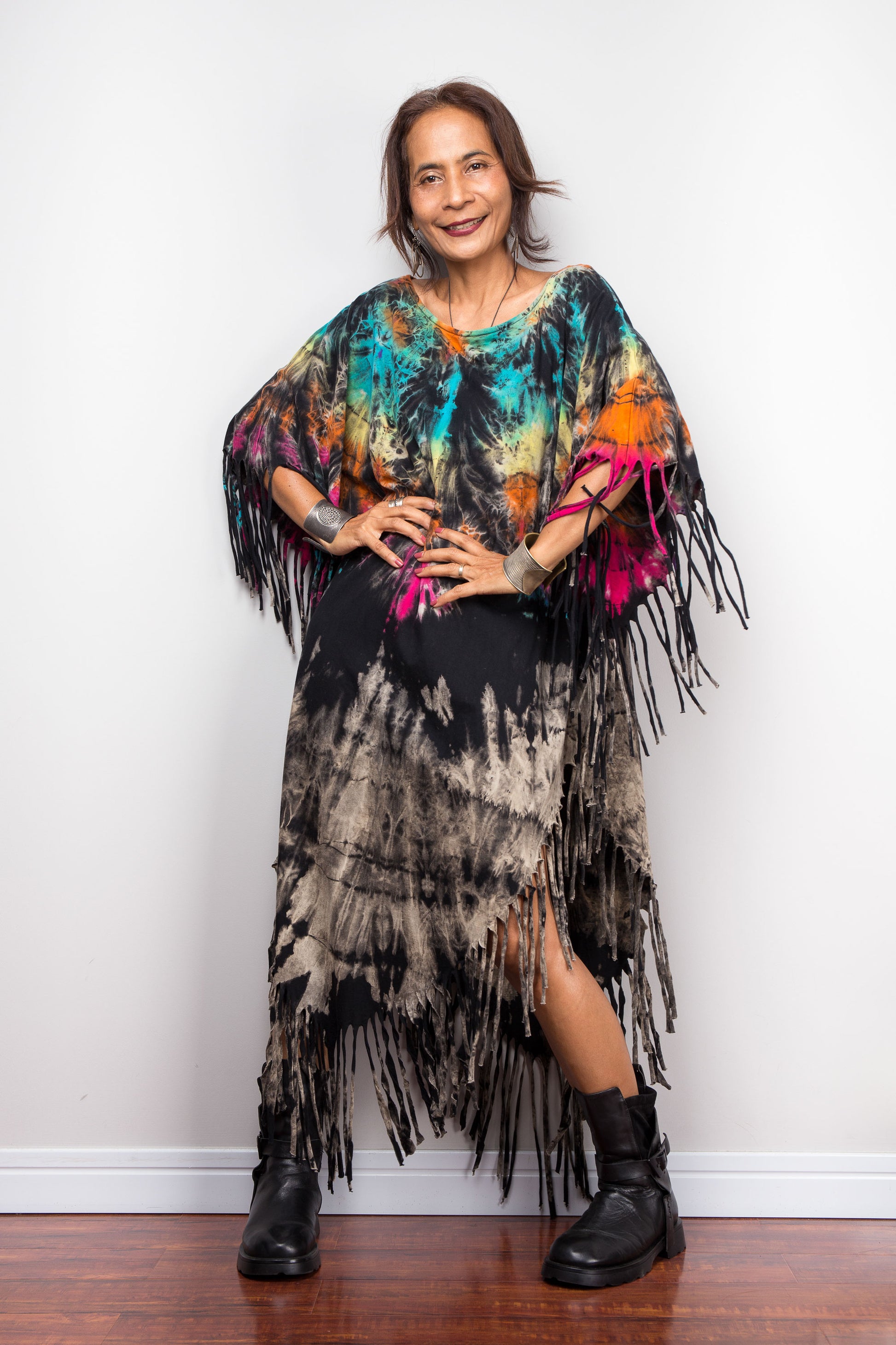 Woman wearing a boho tie dye poncho with fringe standing against a white wall.