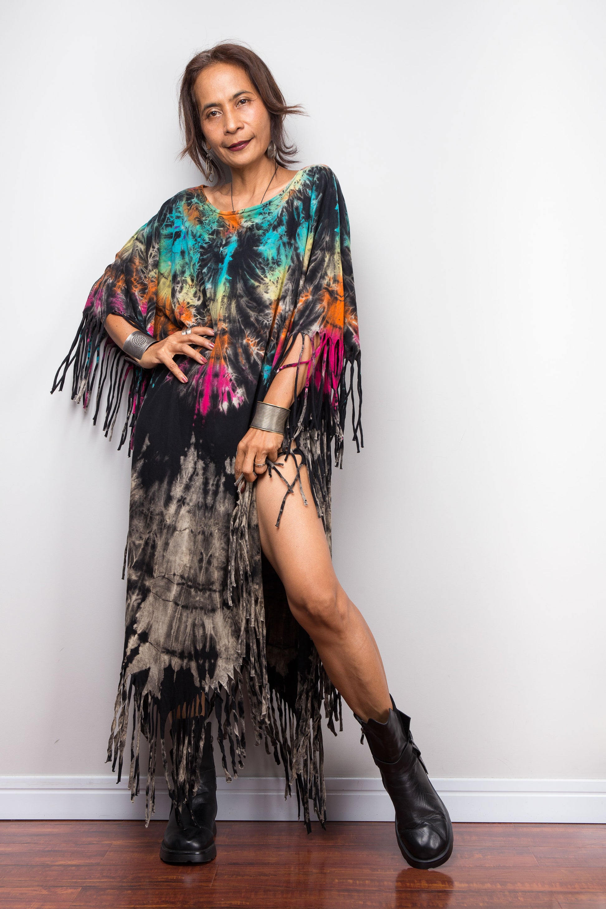 Woman wearing a colorful tie-dye poncho with fringe against a white wall.
