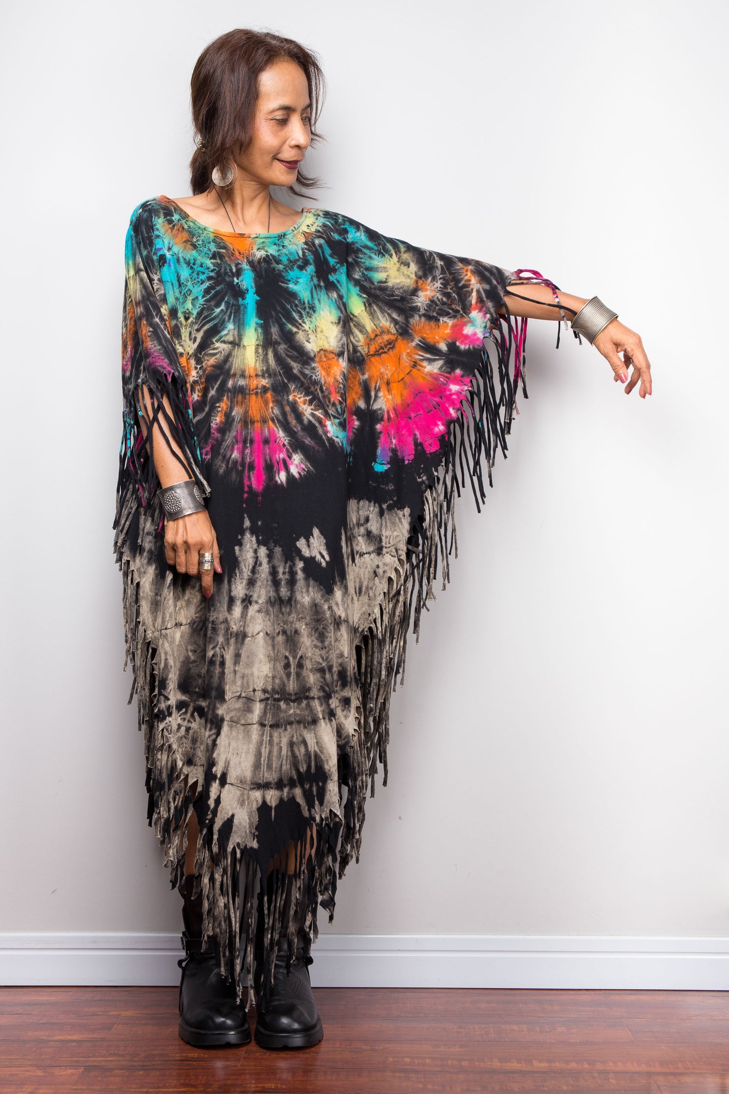 Woman wearing a colorful tie dye poncho tunic dress with fringe against a white wall.