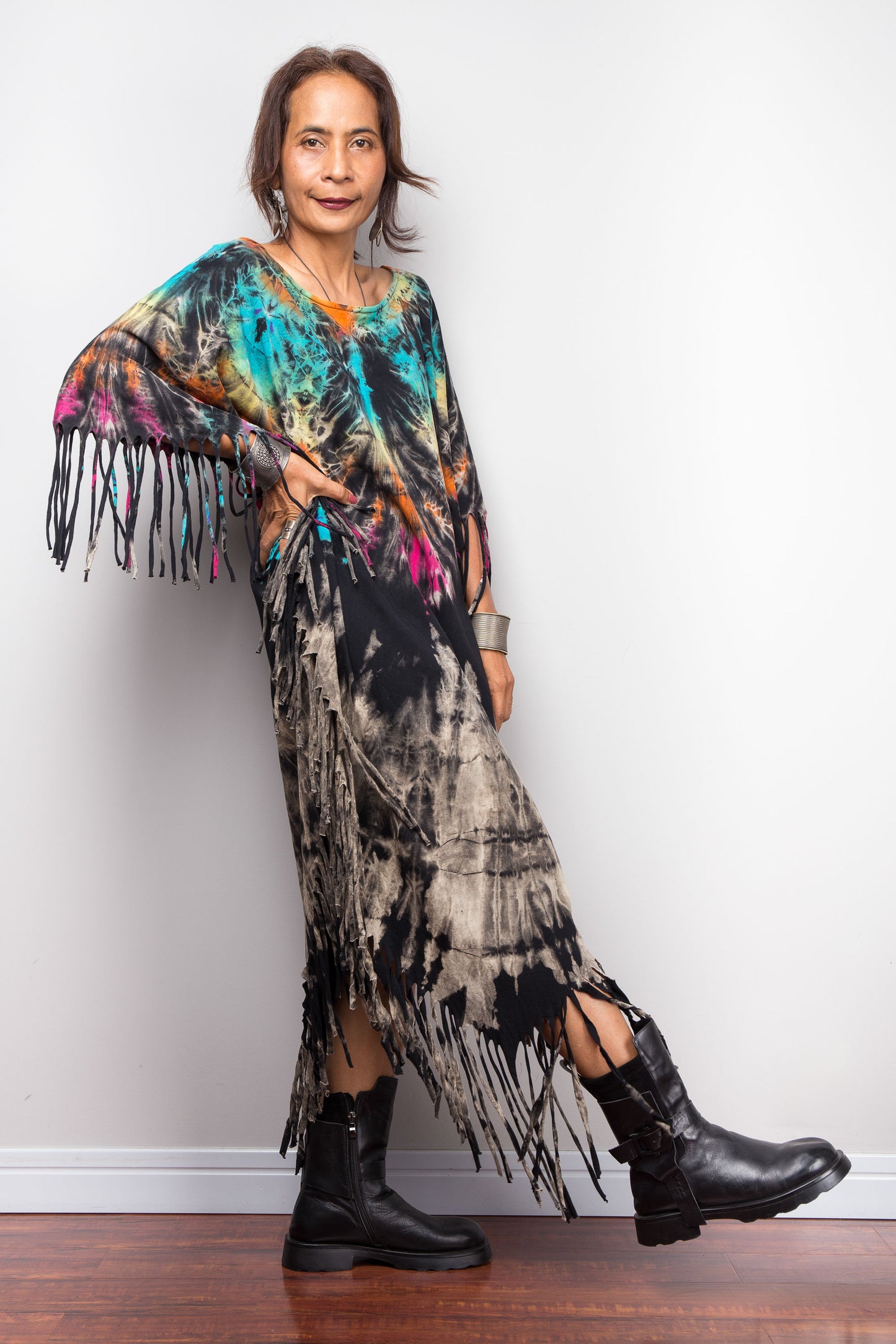 Petite Woman wearing a colorful, fringed tie dye dress and black boots against a white wall.