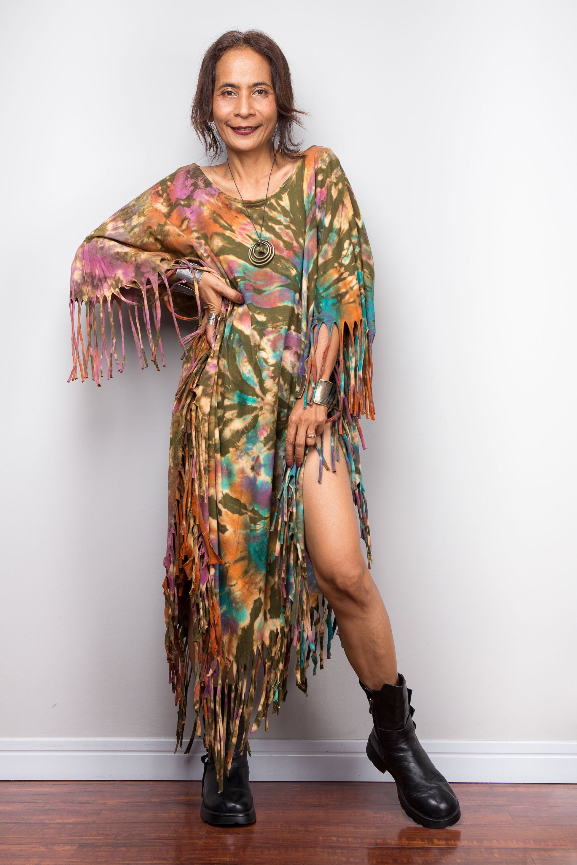 Woman wearing a tie dye poncho dress with fringe and black boots against a white wall.
