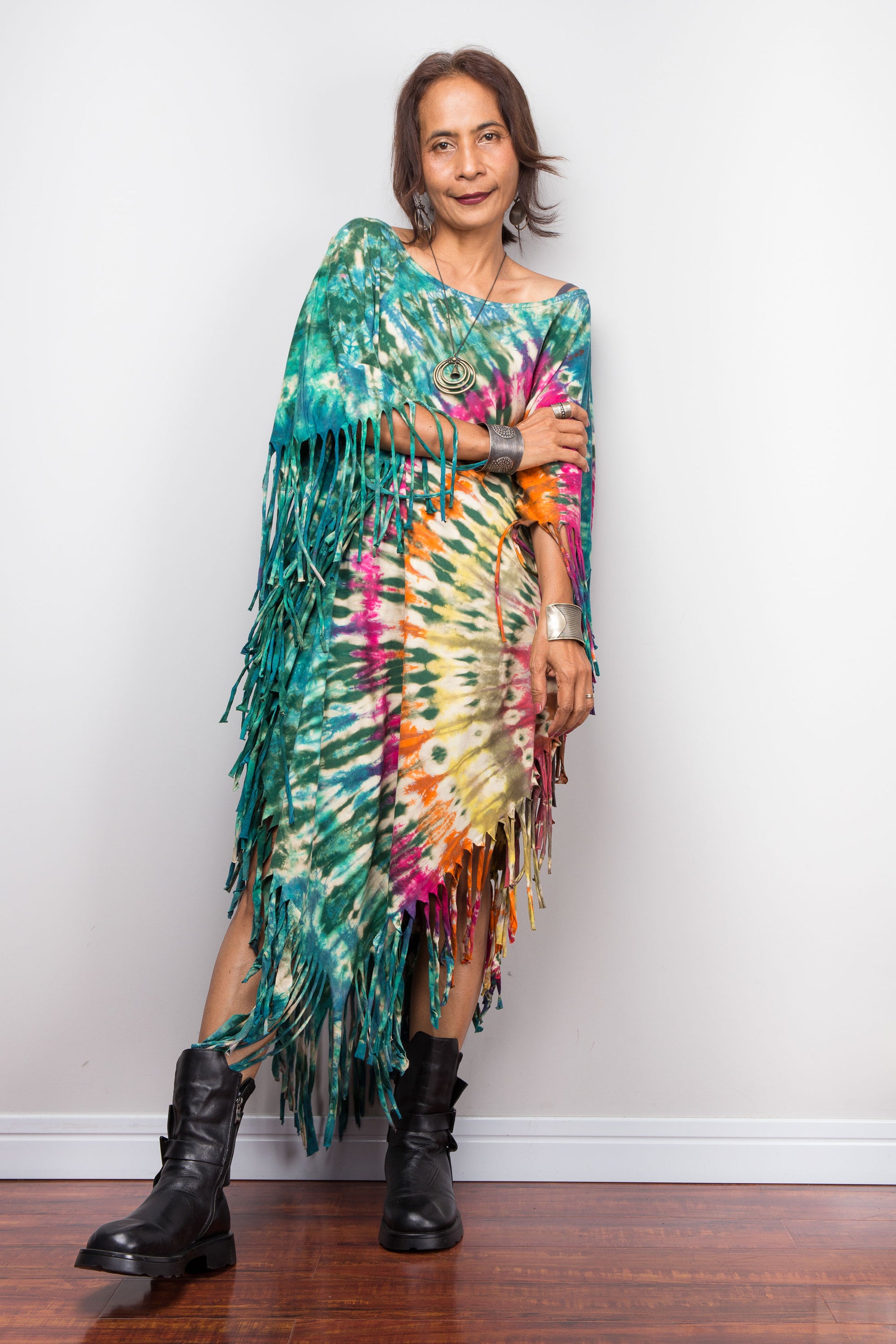 Woman wearing a colorful tie-dye dress with fringe and black boots against a white wall.