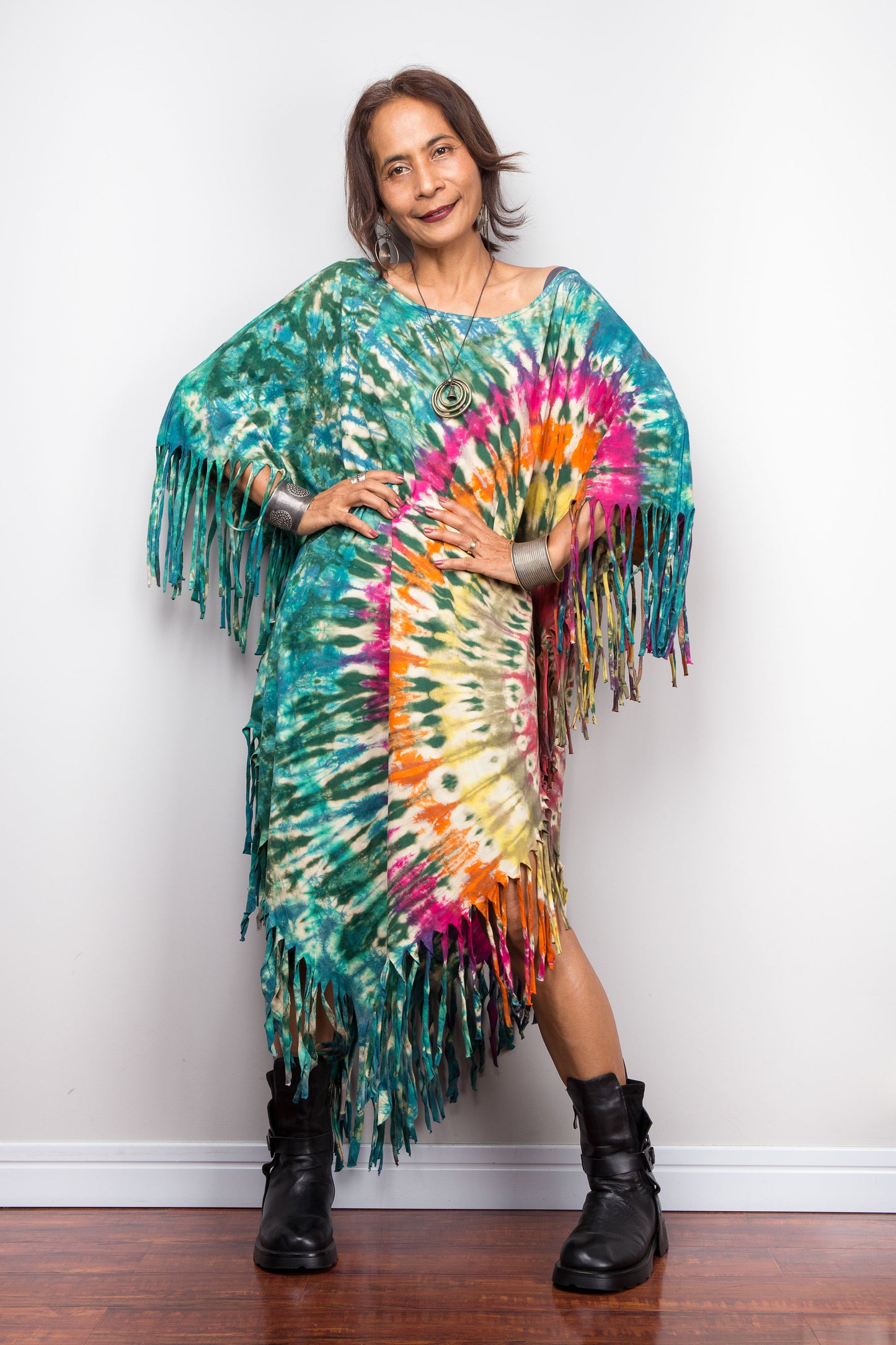 Petite women wearing a colorful tie-dye poncho with fringe against a white wall.