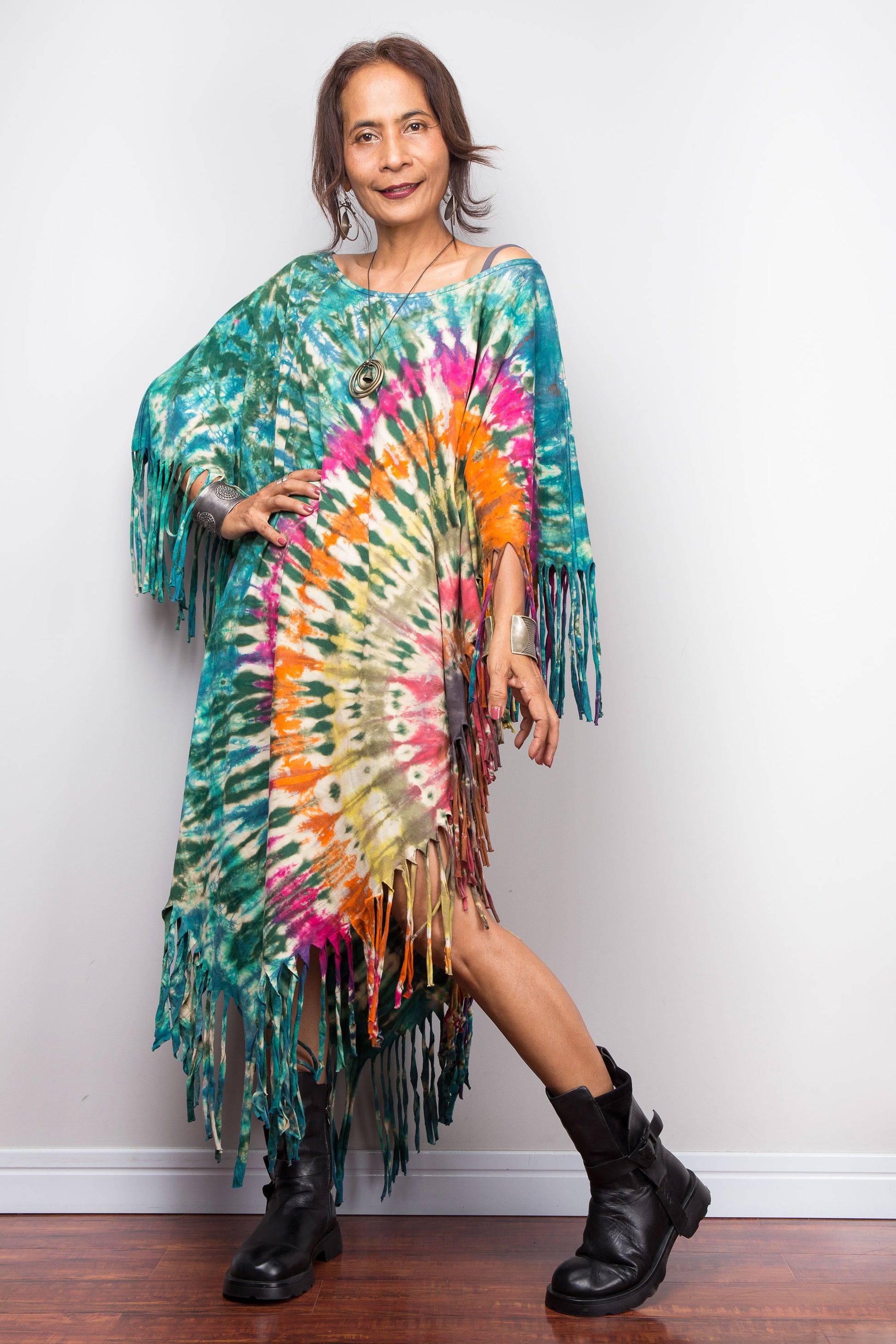 Woman wearing a colorful tie-dye poncho with fringes against a white wall.
