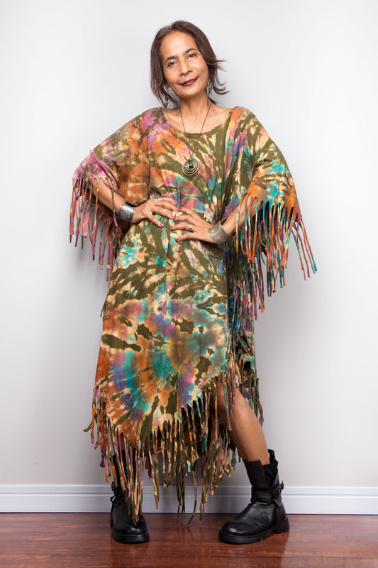 Woman wearing a colorful tie dye poncho dress with fringe details against a white wall.