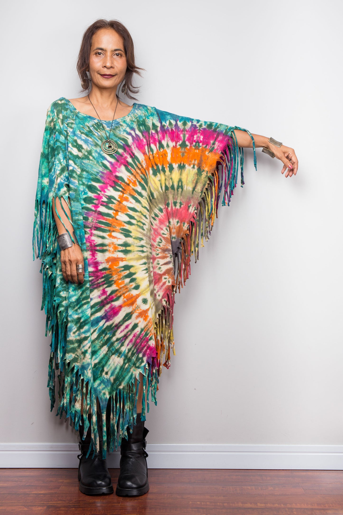 Person wearing a colorful boho tie dye poncho dress with fringes against a white wall.