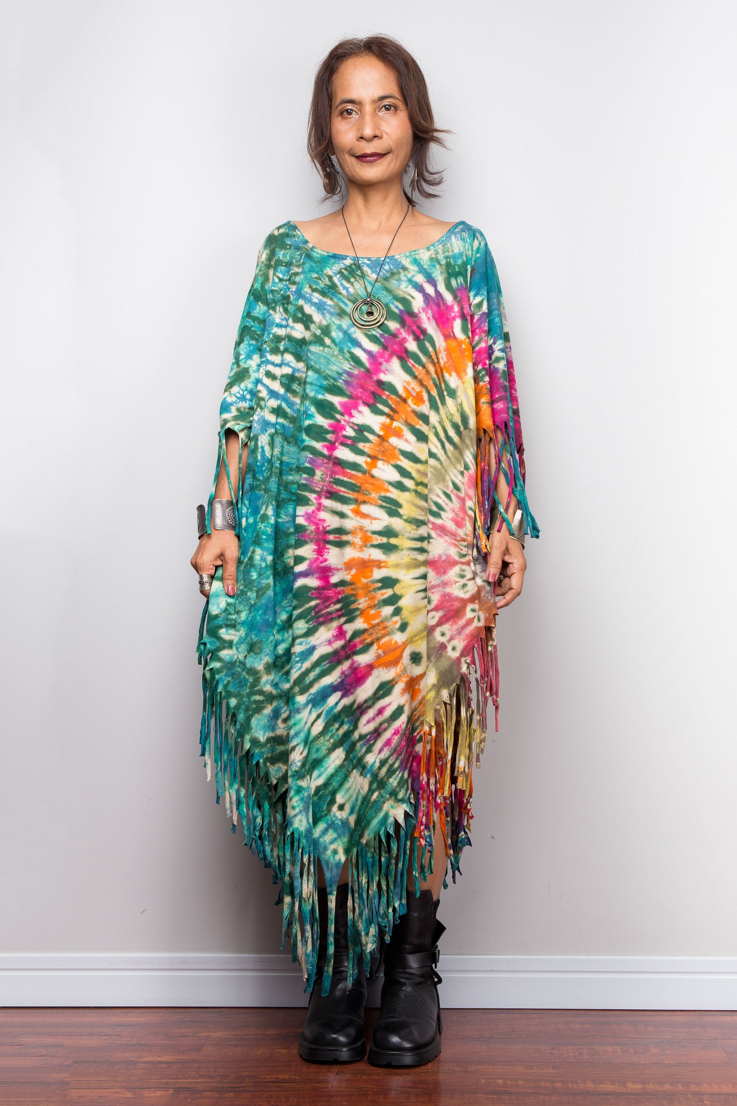 Petite woman wearing a colorful tie dye poncho dress with fringe against a grey wall.
