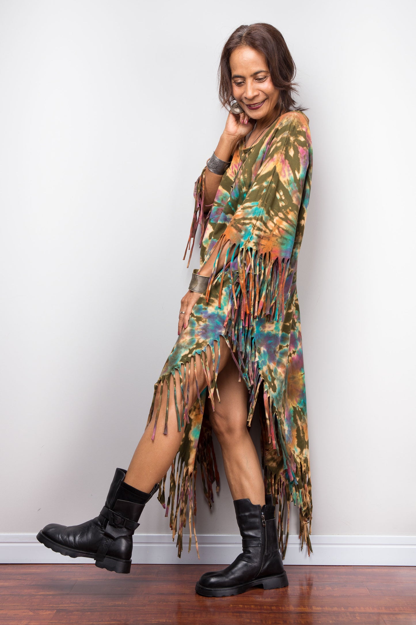 Woman wearing a colorful tie dye fringe dress and black boots against a grey wall.