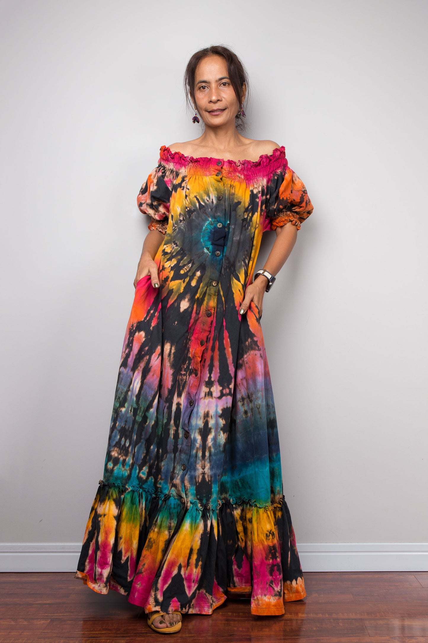 A woman modeling a vibrant, multicolor Nuichan tie-dye maxi dress with puff sleeves and a tiered ruffled hem, standing against a plain light gray wall.