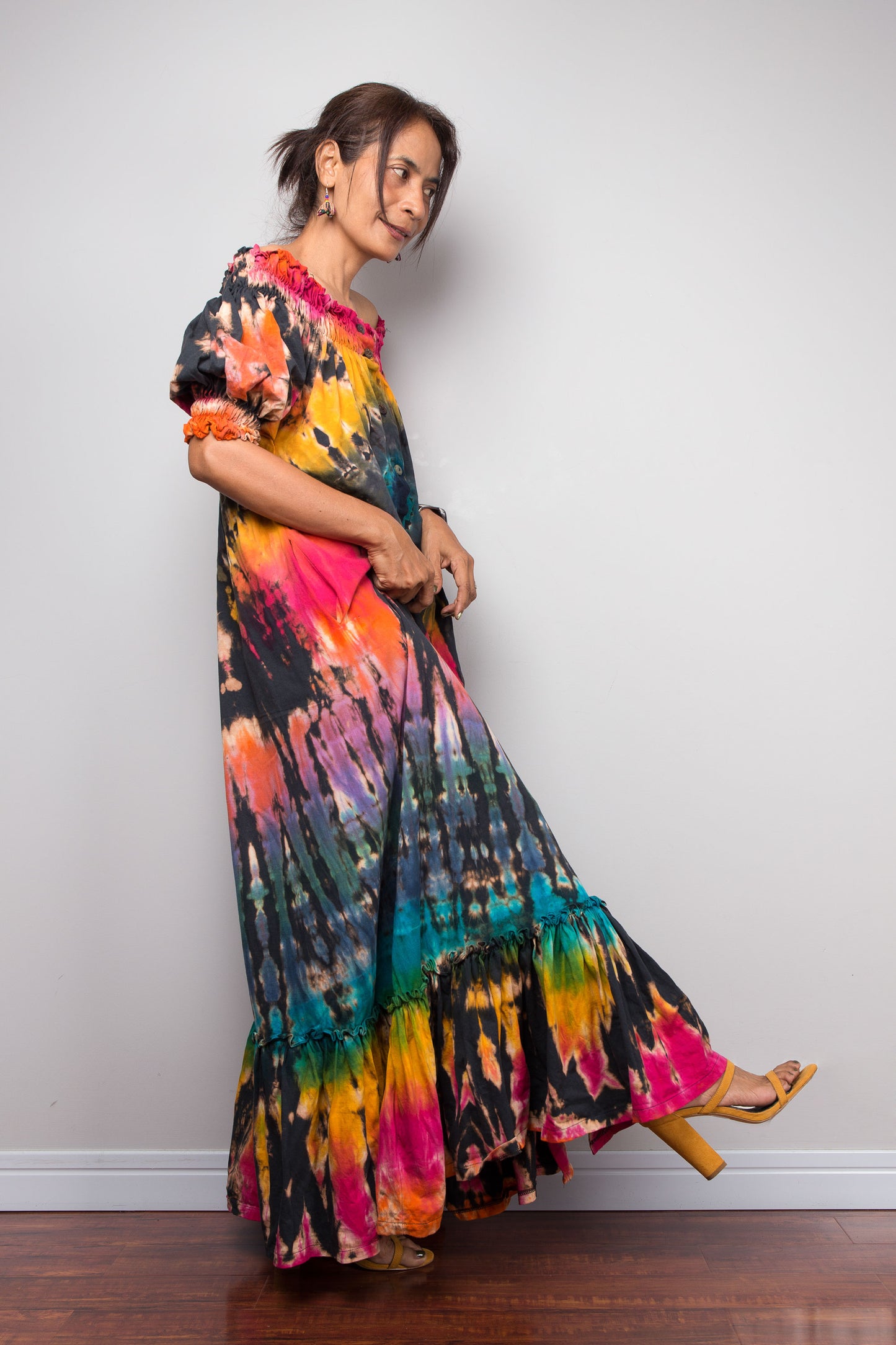 A woman modeling a long, colorful Nuichan tie-dye maxi dress with puff sleeves and a tiered ruffled hem, standing against a plain light gray wall on a dark wood floor.
