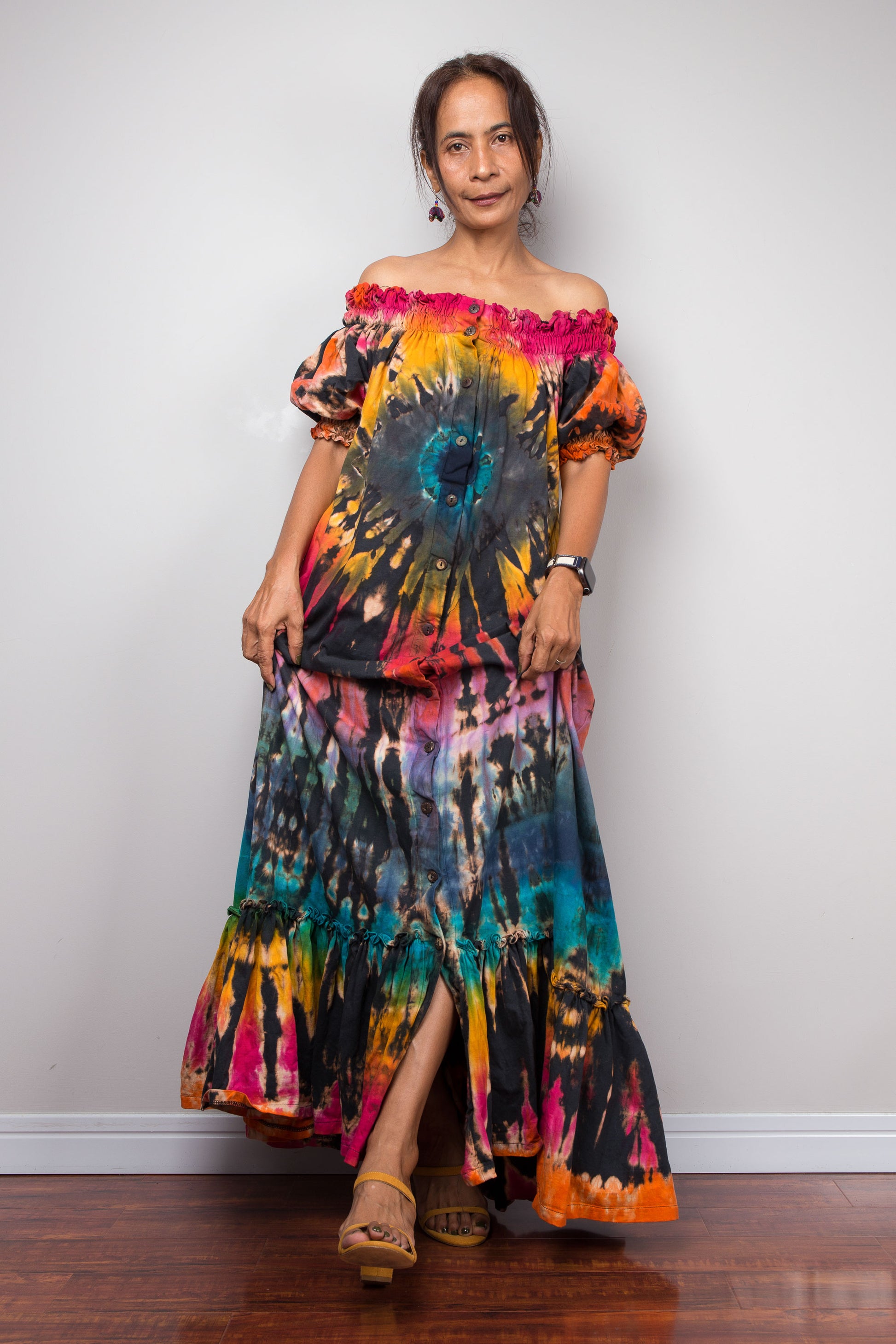 A woman modeling a vibrant tie-dye maxi dress, by nuichan, with puff sleeves and a ruffled neckline, standing against a plain light gray wall on a wooden floor.